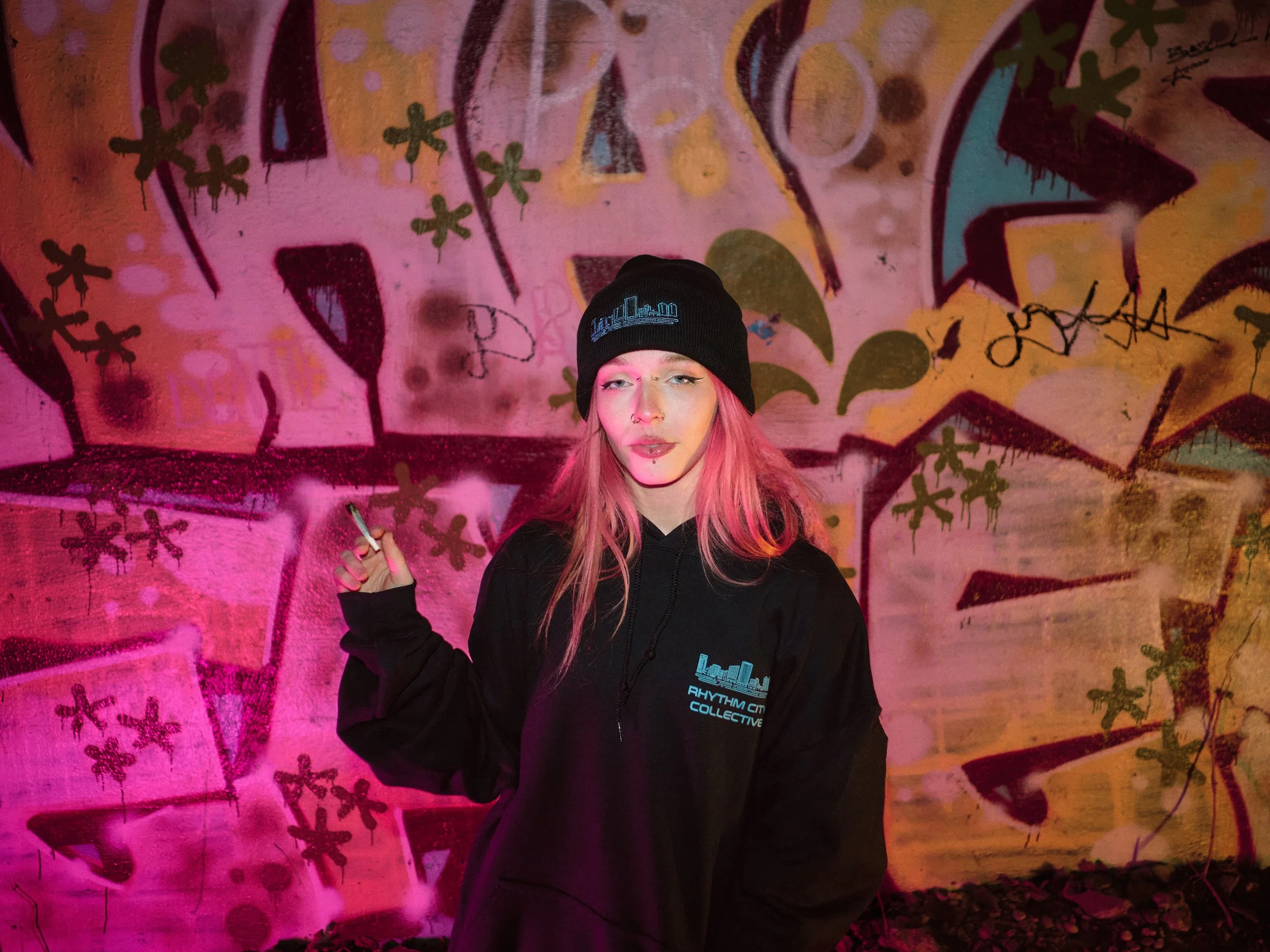 A young woman with pink hair, wearing a black beanie and a black hoodie with a city skyline and text that reads 'Rhythm City Collective'. She is standing in front of a colorful graffiti wall illuminated with pink and purple lighting, holding a cigarette in her right hand.