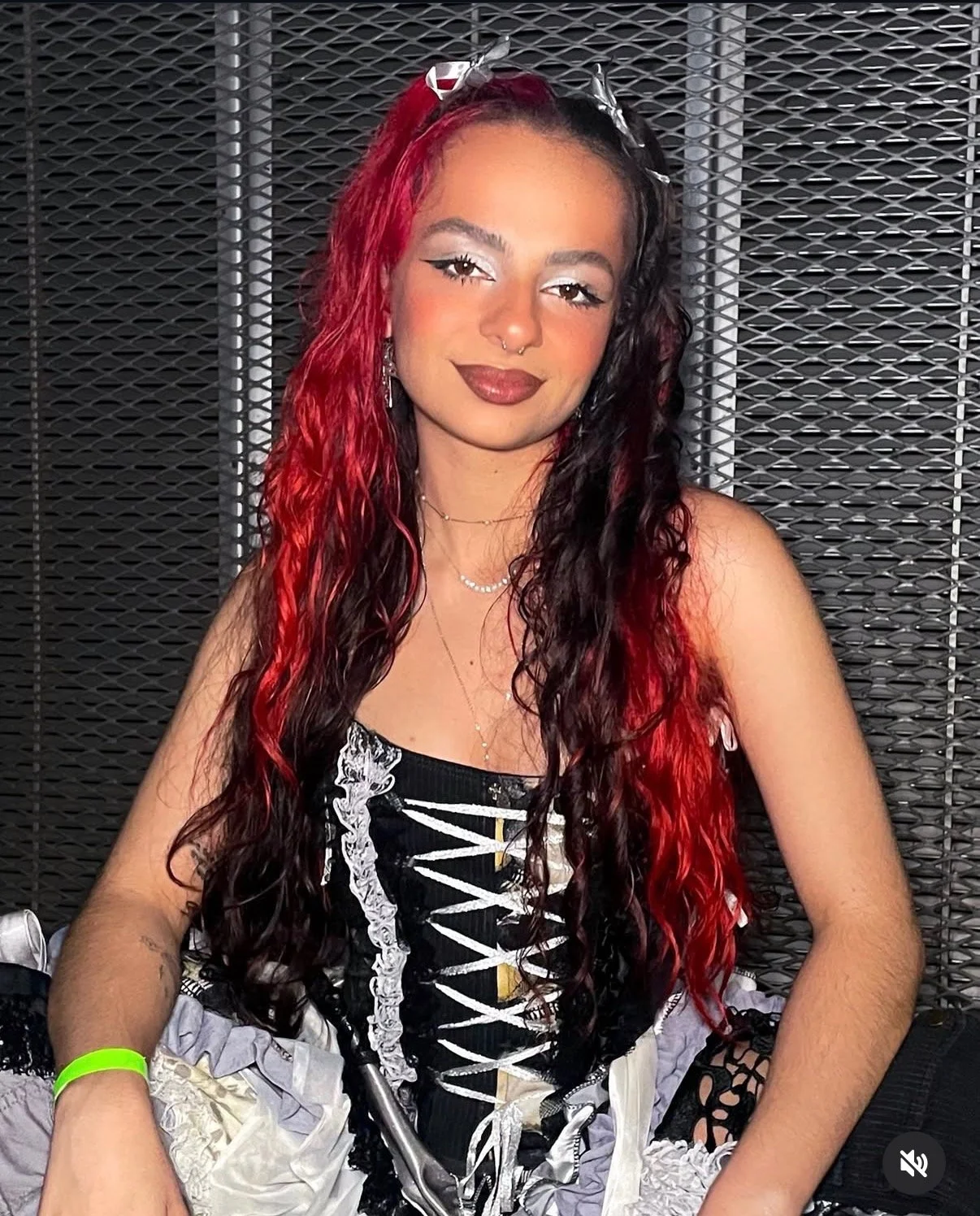 A woman with long, wavy, half-black and half-red hair, wearing a black and white corset and matching skirt, sitting against a metallic backdrop.