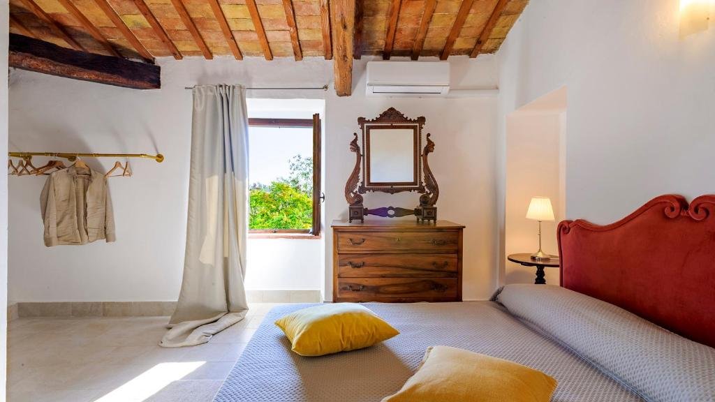 A bedroom with a wooden ceiling and white walls, featuring a red upholstered headboard, a wooden dresser with an ornate mirror, a small nightstand with a lamp, a window with curtains showing green trees outside, and a coat rack with beige clothing hanging.