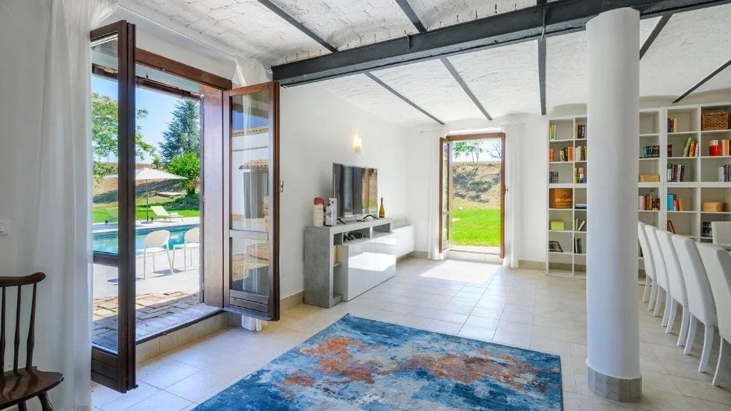 Bright living room with open balcony door leading to a patio with outdoor seating and a swimming pool. The room has white walls, a large blue and orange rug, a TV on a stand, a white bookshelf filled with books, and white dining chairs along a table.