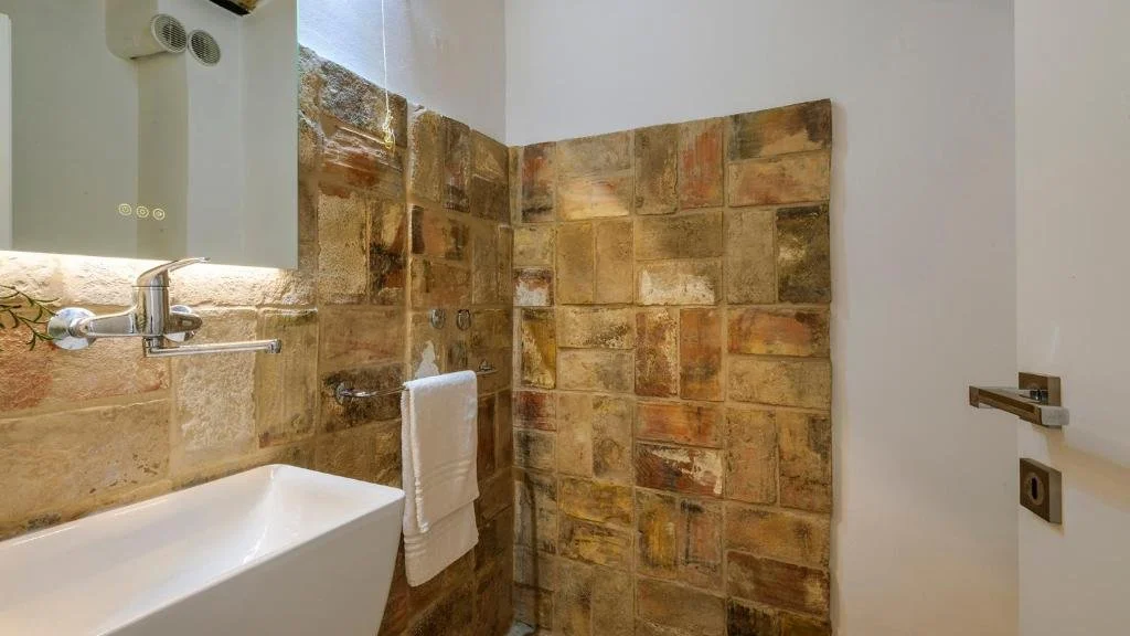 Small bathroom with a white sink, a mirror, and a partially tiled stone wall in the shower area, with a towel hanging on a hook.