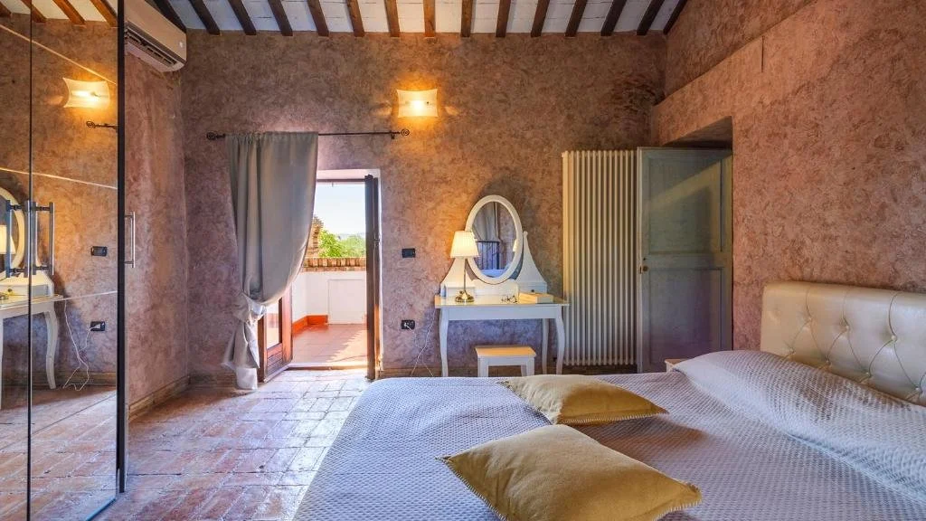 A bedroom with an open balcony door, a bed with beige pillows, a vanity with a mirror, and wall-mounted lamps.