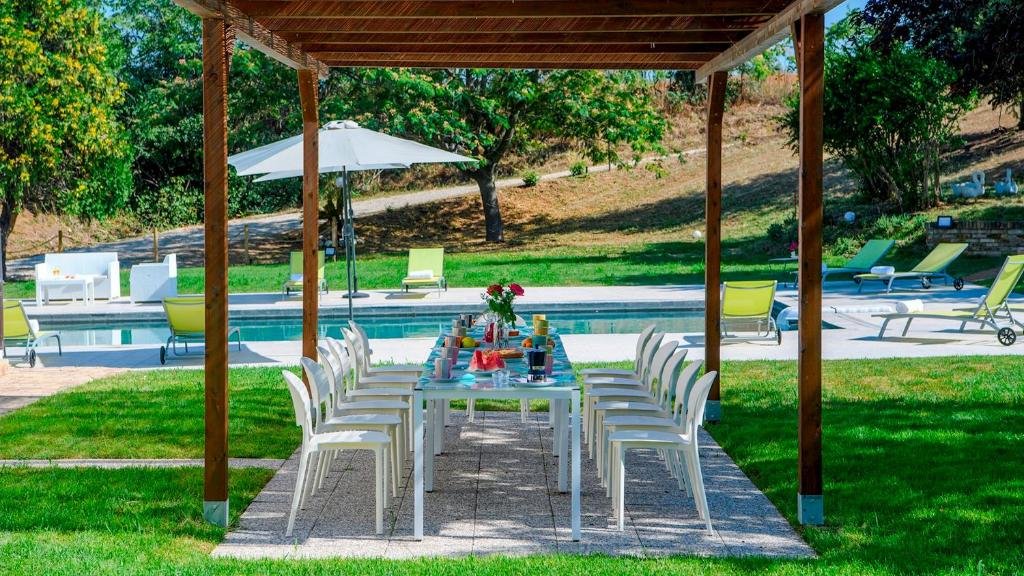Outdoor patio with a dining table and chairs under a wooden pergola, pool in the background with lounge chairs, umbrellas, and green trees.