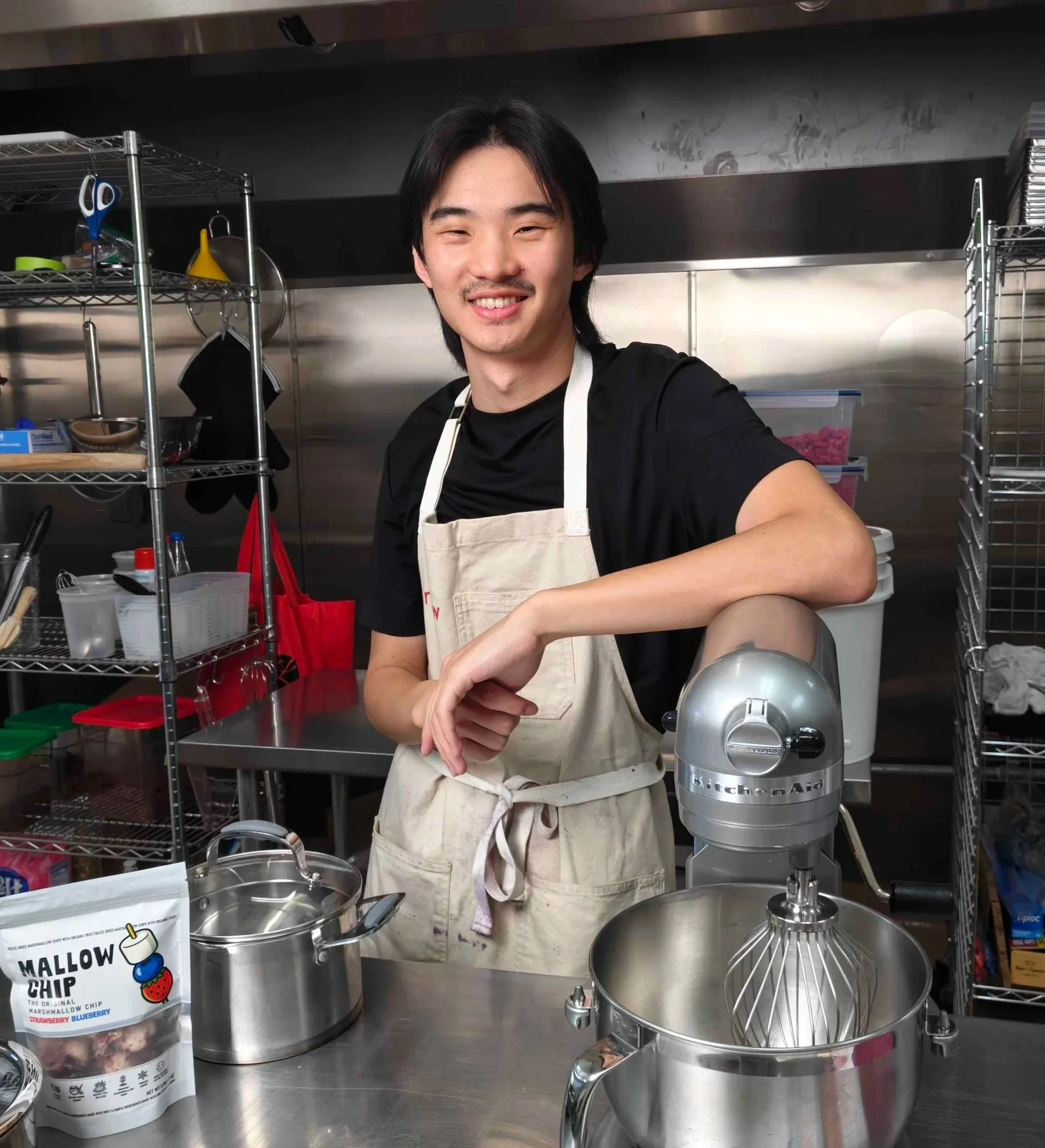 A smiling man wearing a black shirt and a white apron standing in a professional kitchen with stainless steel shelves and cooking equipment, including a KitchenAid mixer and labelling for muffin or cupcake batter with a cartoon marshmallow and berry theme.