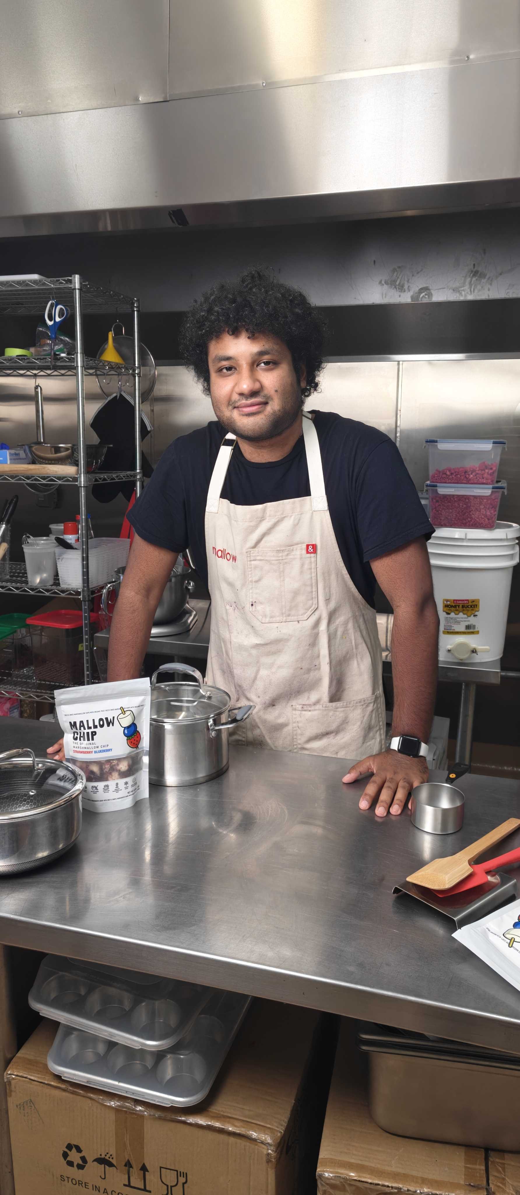 A man with black curly hair and a beard stands in a commercial kitchen, wearing a beige apron over a black T-shirt, smiling. There are kitchen utensils, shelves with containers, and a knife holder visible behind him. On the counter in front, there are cooking tools, a package of marshmallow chips, a metal pot, and a small wooden spatula.