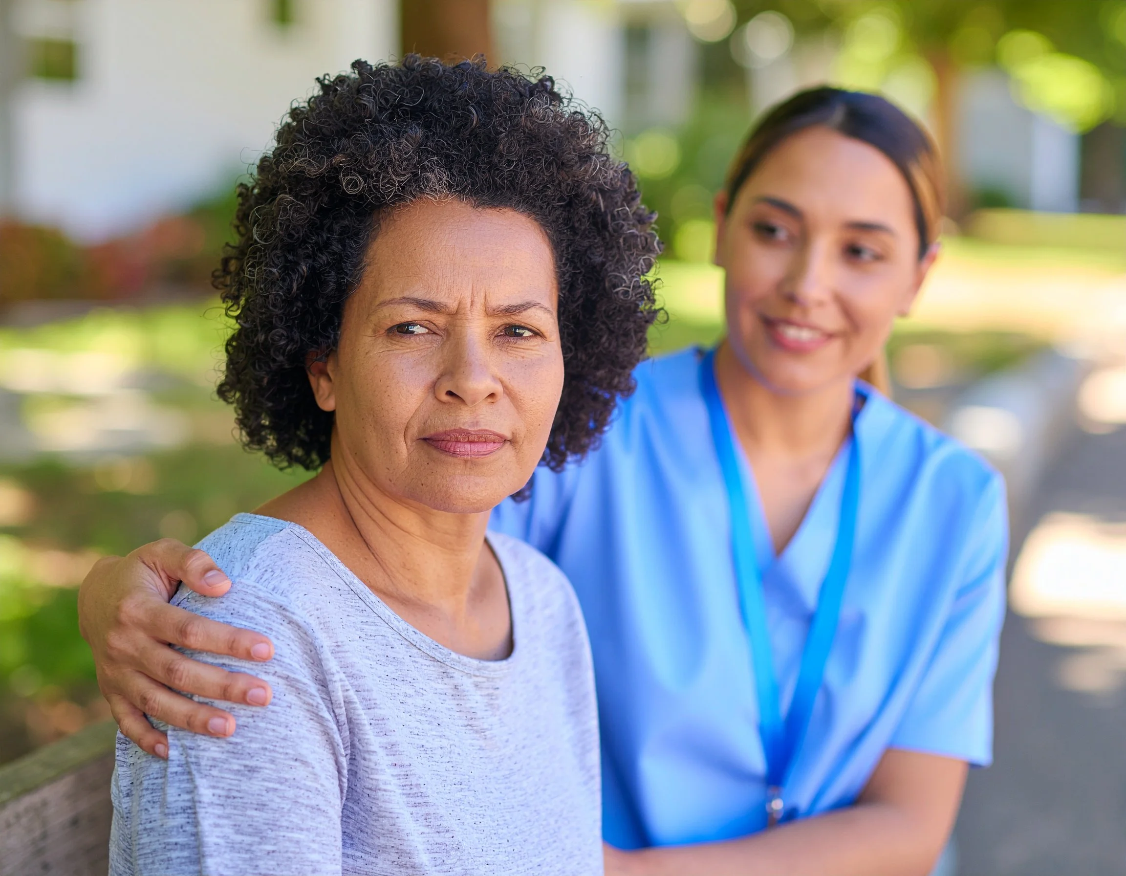 A woman with curly dark hair and a serious expression is sitting on a park bench, with a healthcare worker in blue scrubs and a name badge next to her, smiling. The healthcare worker has her hand on the woman's shoulder in a supportive gesture. They are outdoors with green trees and sunlight in the background.
