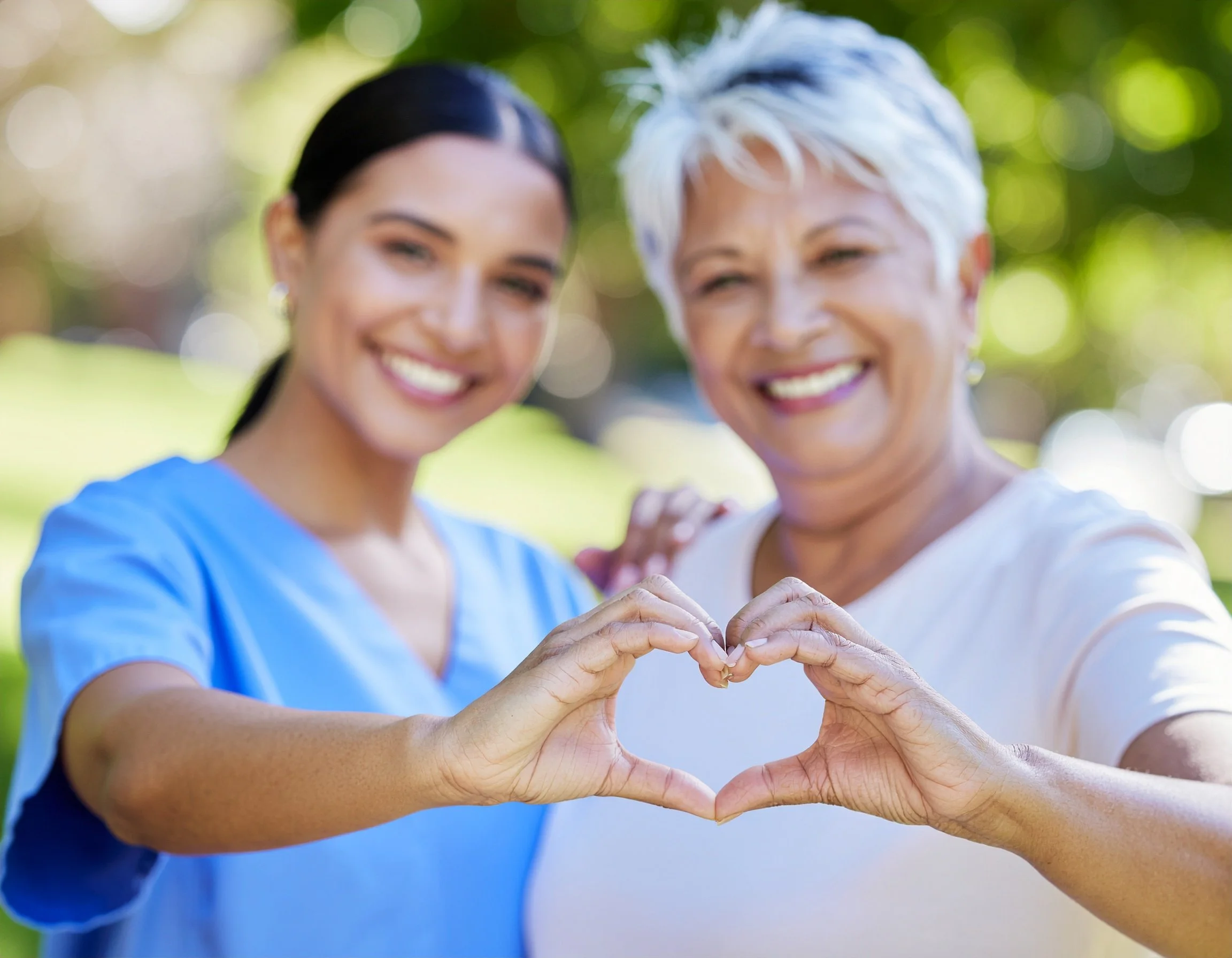 Two women, one younger and one older, smiling outside and forming a heart with their hands.
