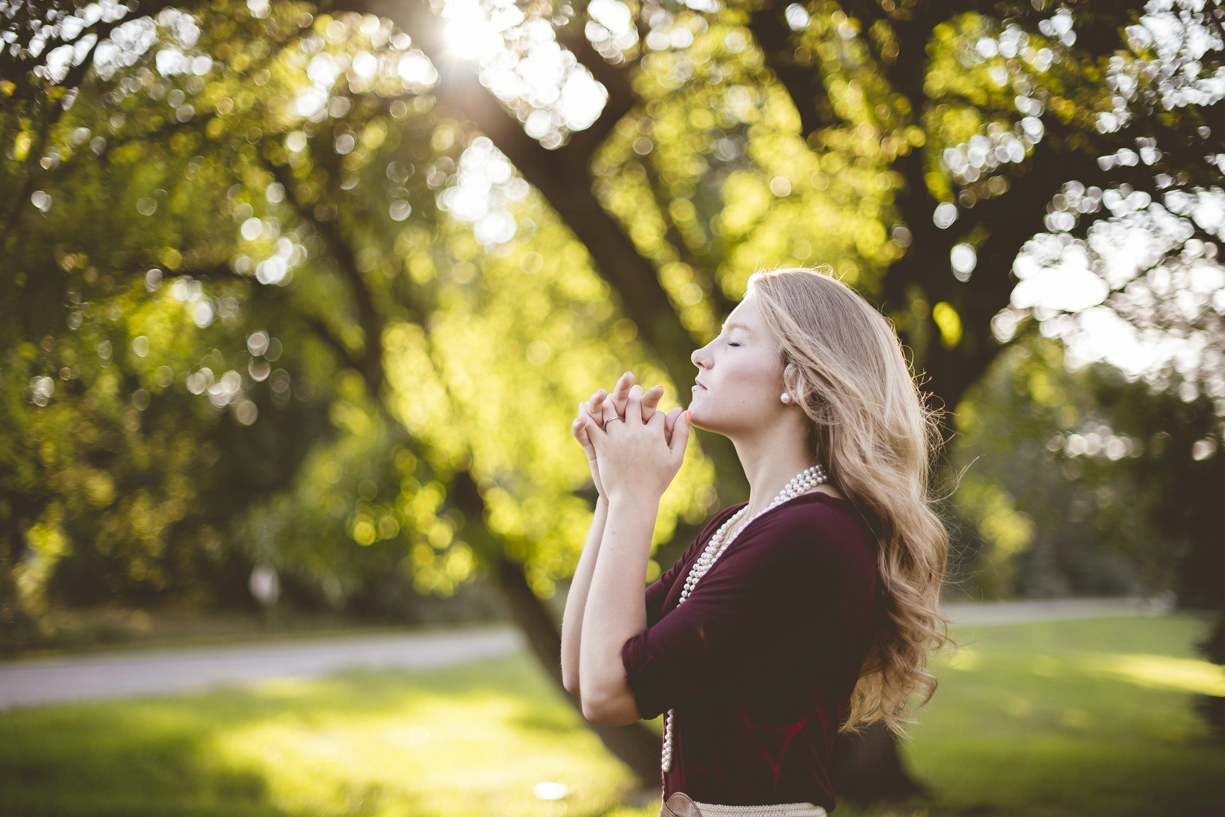 A woman with long, wavy blonde hair and pearl earrings is standing outdoors with her eyes closed and her hands clasped together near her chin. She is wearing a dark red dress and a pearl necklace. The background shows green trees and sunlight filtering through the leaves.
