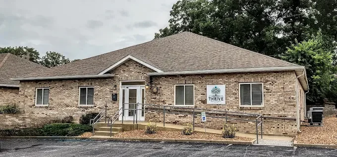 Single-story brick building with a gabled roof, a white entrance door, four windows, and a sign that says 'THRIVE' with a tree logo, surrounded by a small landscaped area and parking lot.