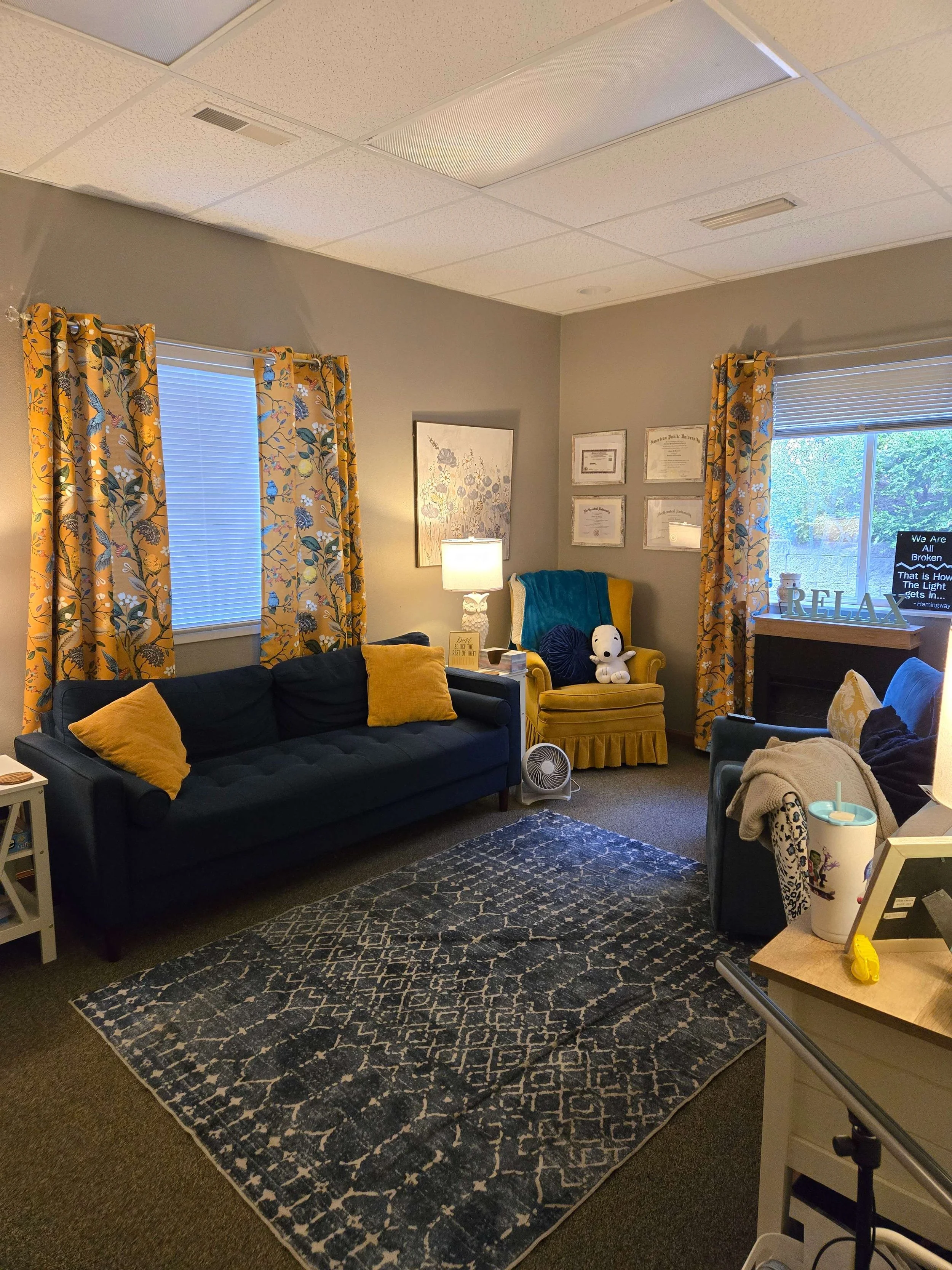 A cozy living room with a navy blue sofa, yellow pillows, and matching curtains. There is a yellow armchair with a Snoopy plush toy, a small white fan, and a side table with a drink cup. The room has framed certificates or artwork on the wall, a lamp, a rug, and a window bringing in natural light.