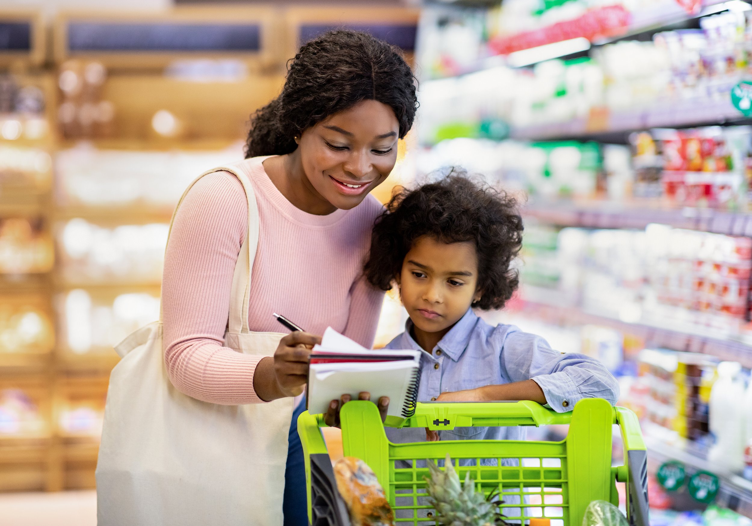 A woman and a young girl shopping in a grocery store, examining a notepad together over a green shopping cart with fruits like pineapples and papaya inside.