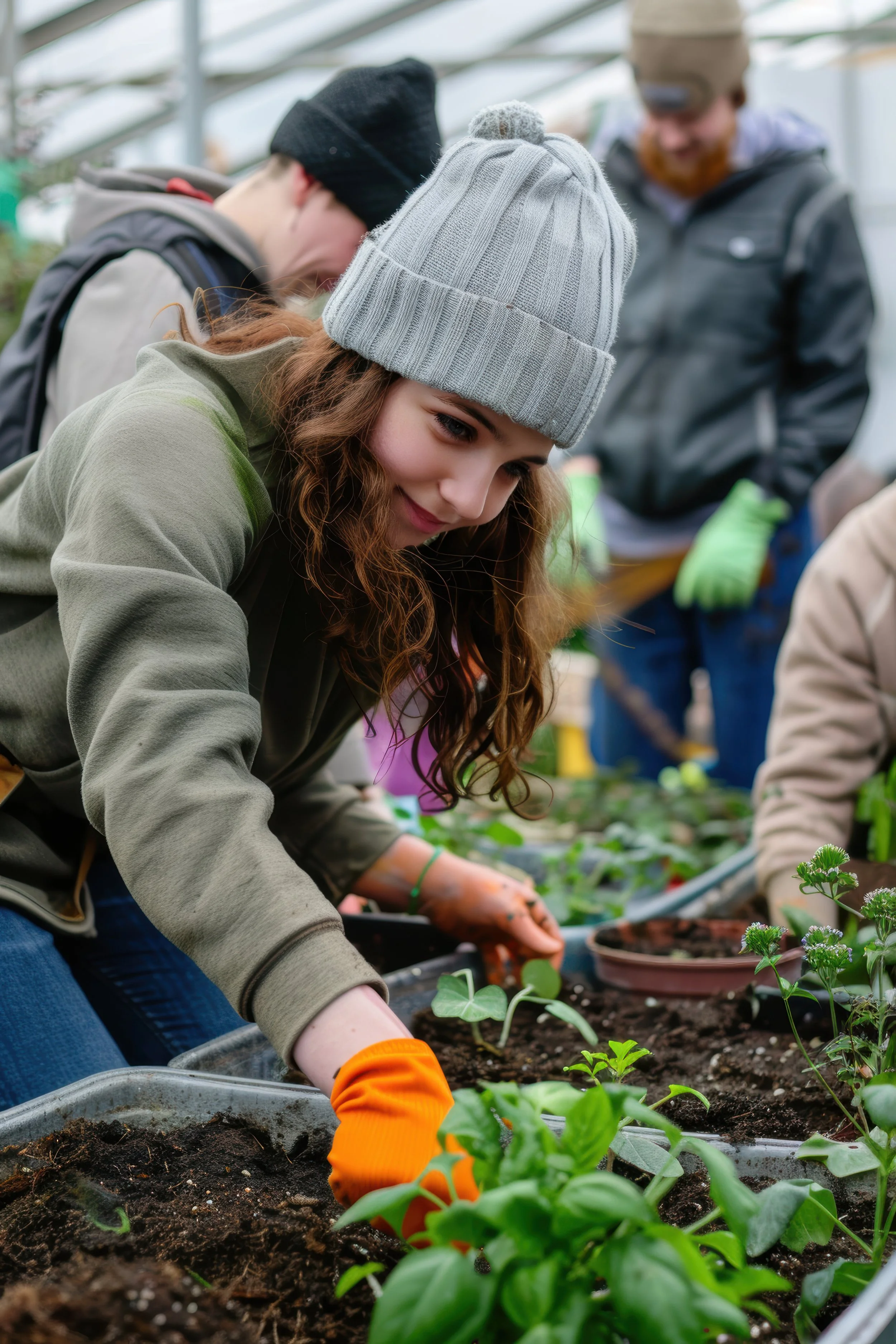 Young woman in a gray beanie and orange gardening gloves planting seedlings in a greenhouse with other people working in the background.