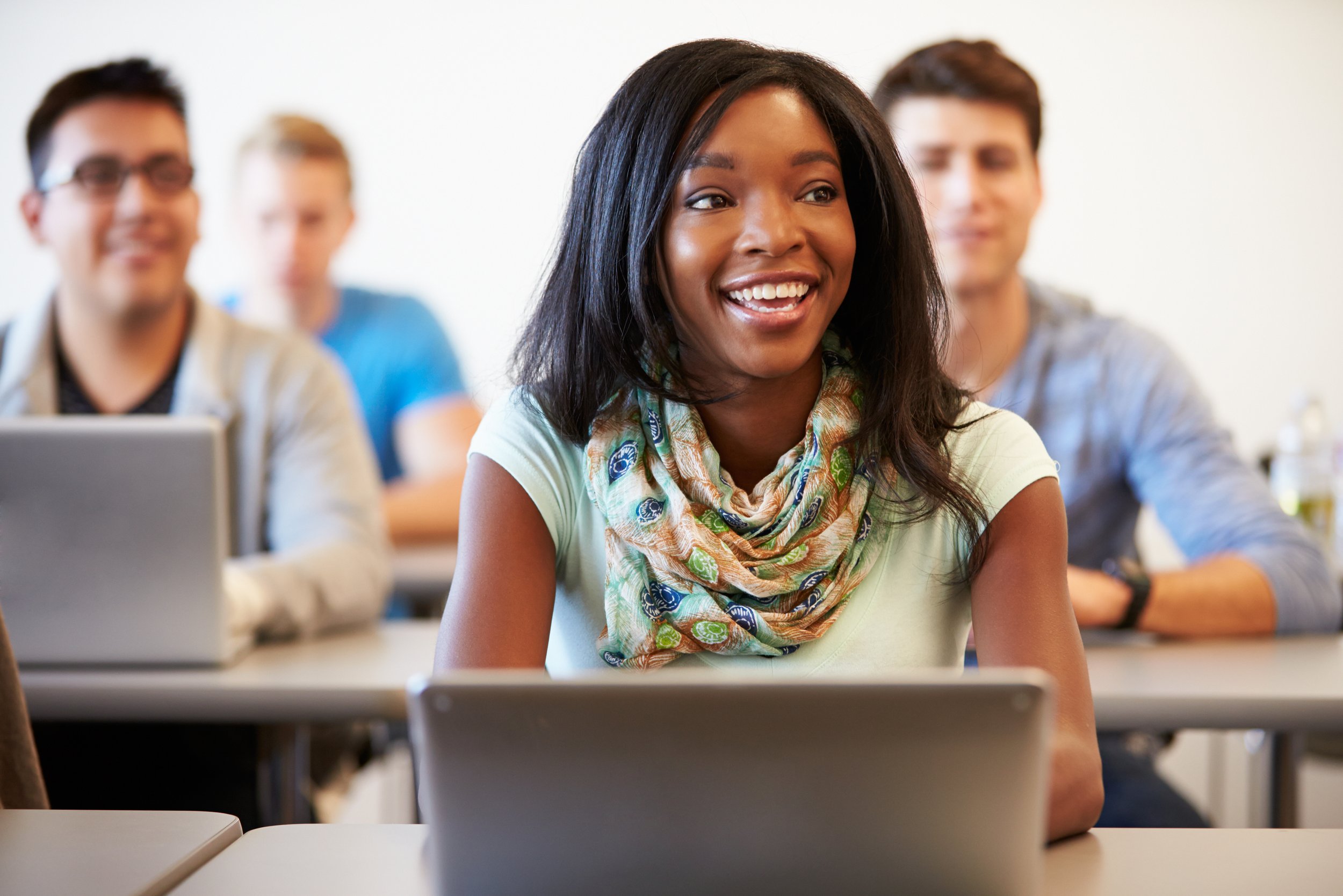 A woman smiling in a classroom or meeting room with four other students or attendees in the background, sitting at desks with laptops.