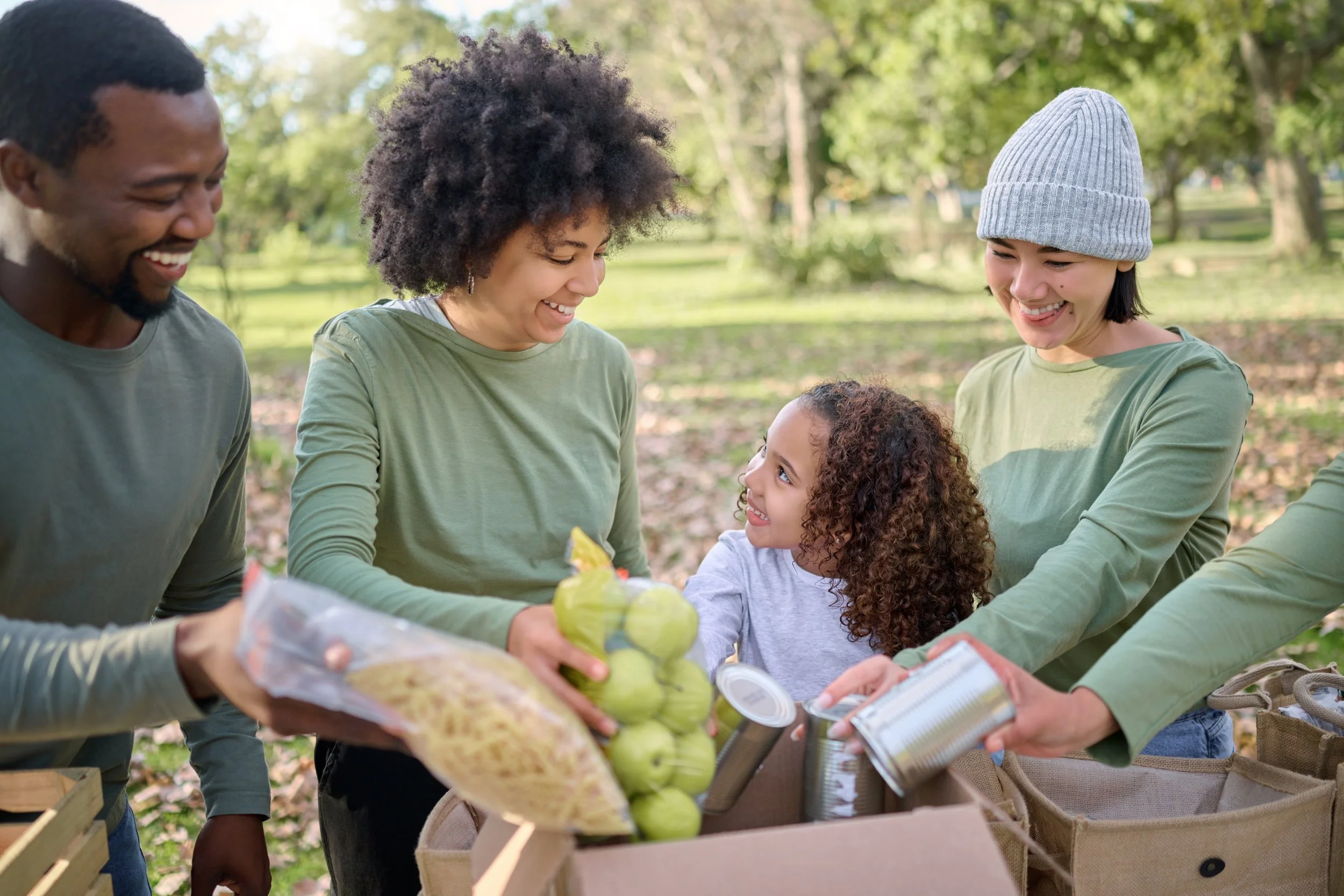 A diverse group of people, including two women, one man, and a young girl, are smiling and sharing groceries outdoors in a park with green trees in the background.