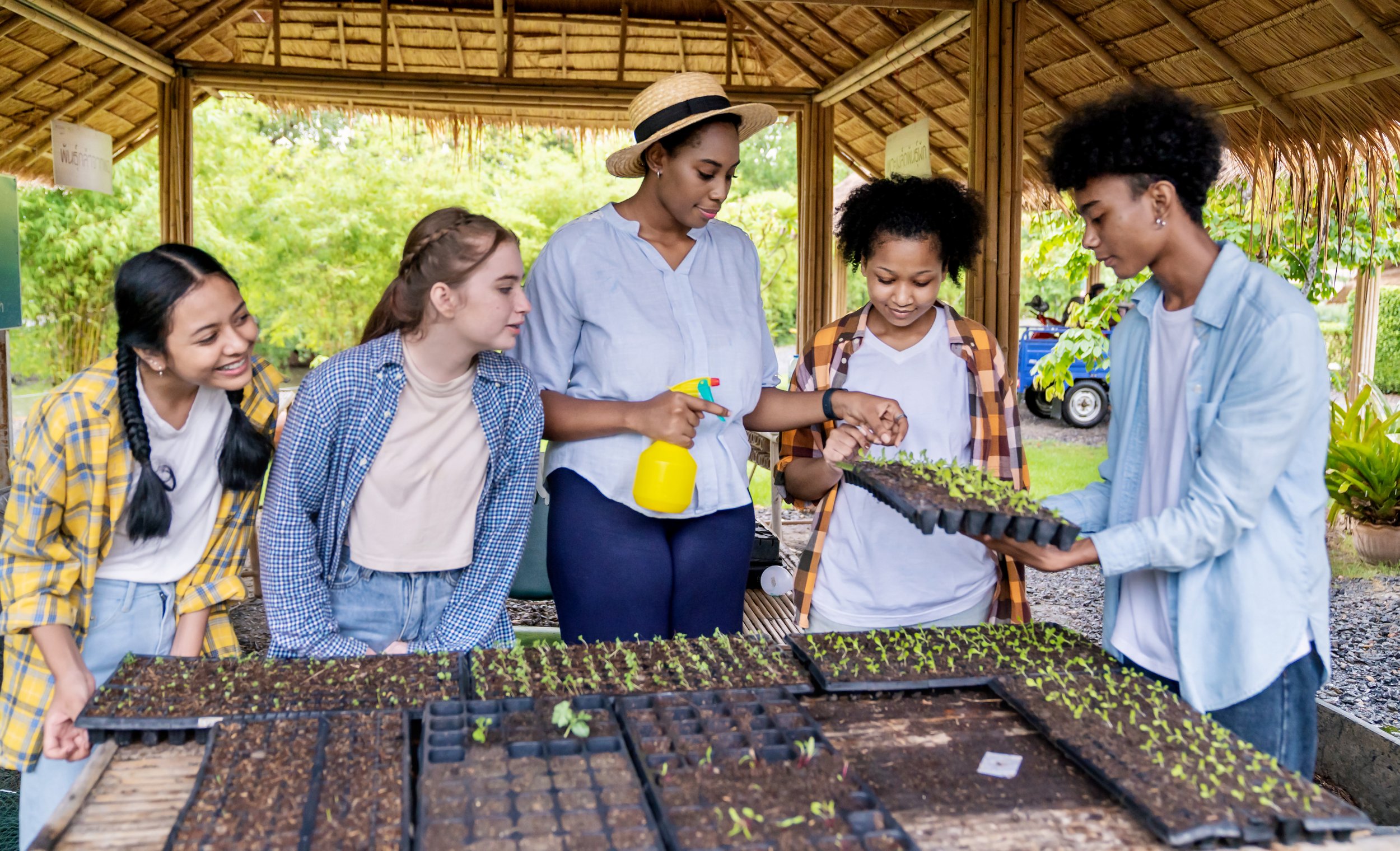 Group of five young women engaging in planting seedlings at a gardening station outdoors, with a thatched roof structure and lush greenery in the background.