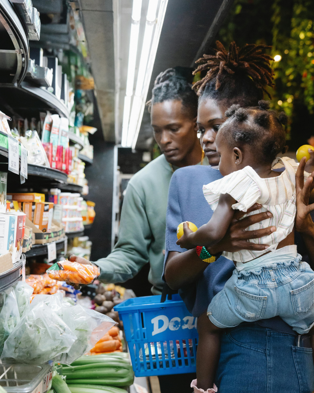 A family shopping for groceries in a store aisle, with a woman holding a young girl and a man selecting vegetables.