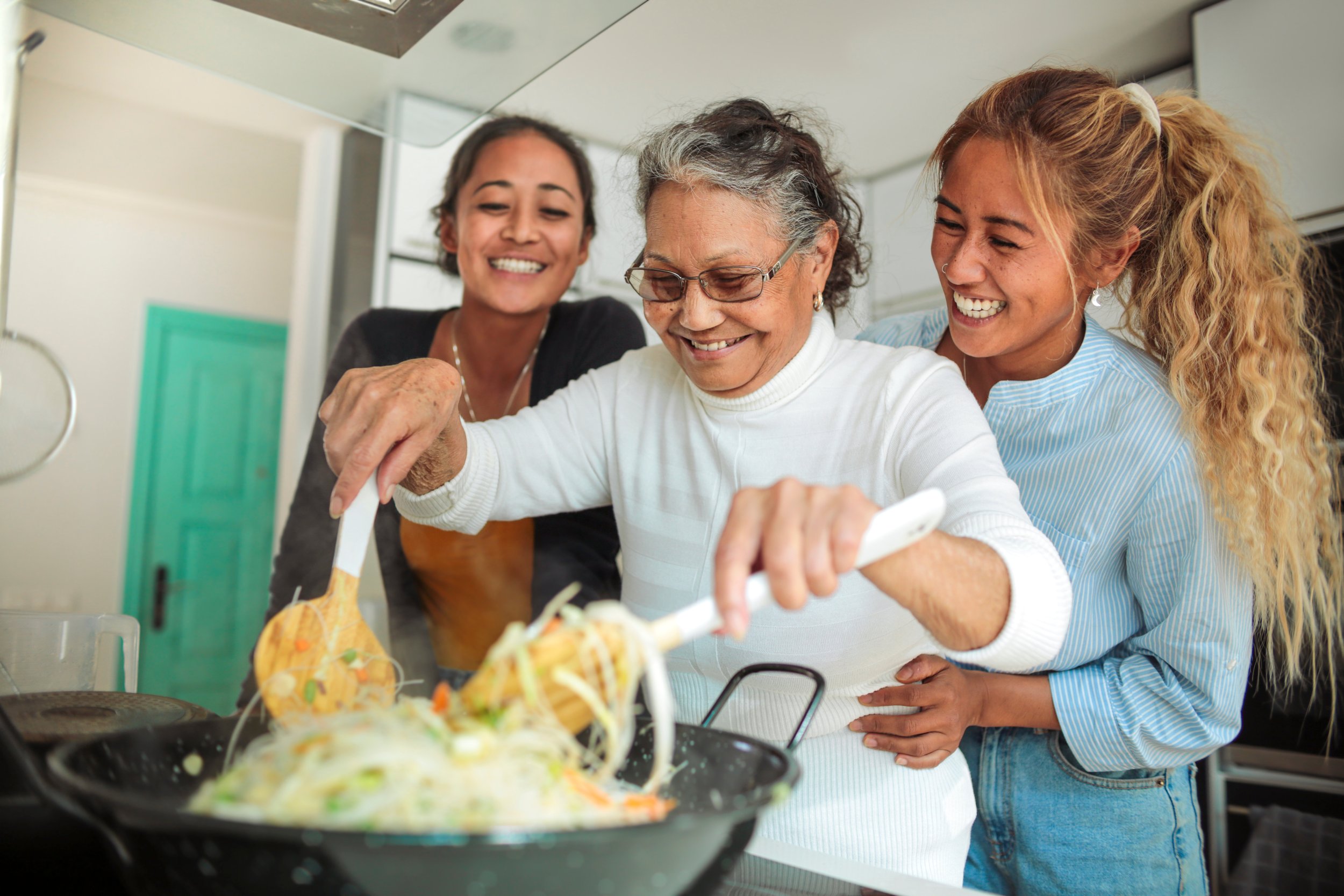 Three women enjoying cooking and sharing a meal in the kitchen, with one woman stirring a pan of food on the stove while the others watch and smile.