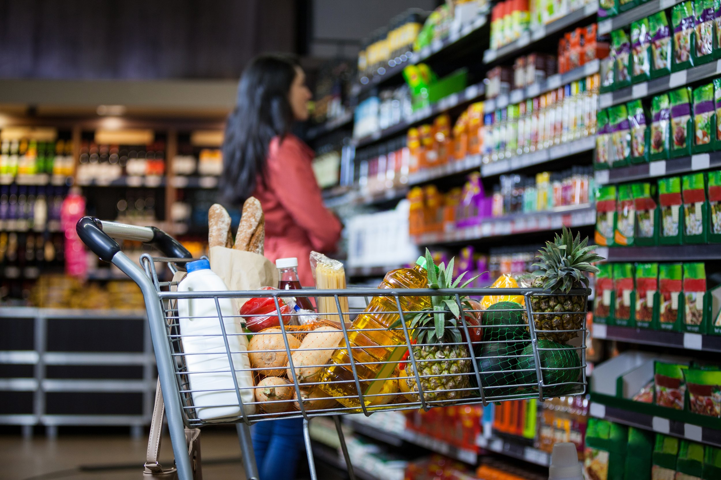 A shopping cart filled with fruits, vegetables, and groceries, parked in a supermarket aisle with a woman shopping in the background.