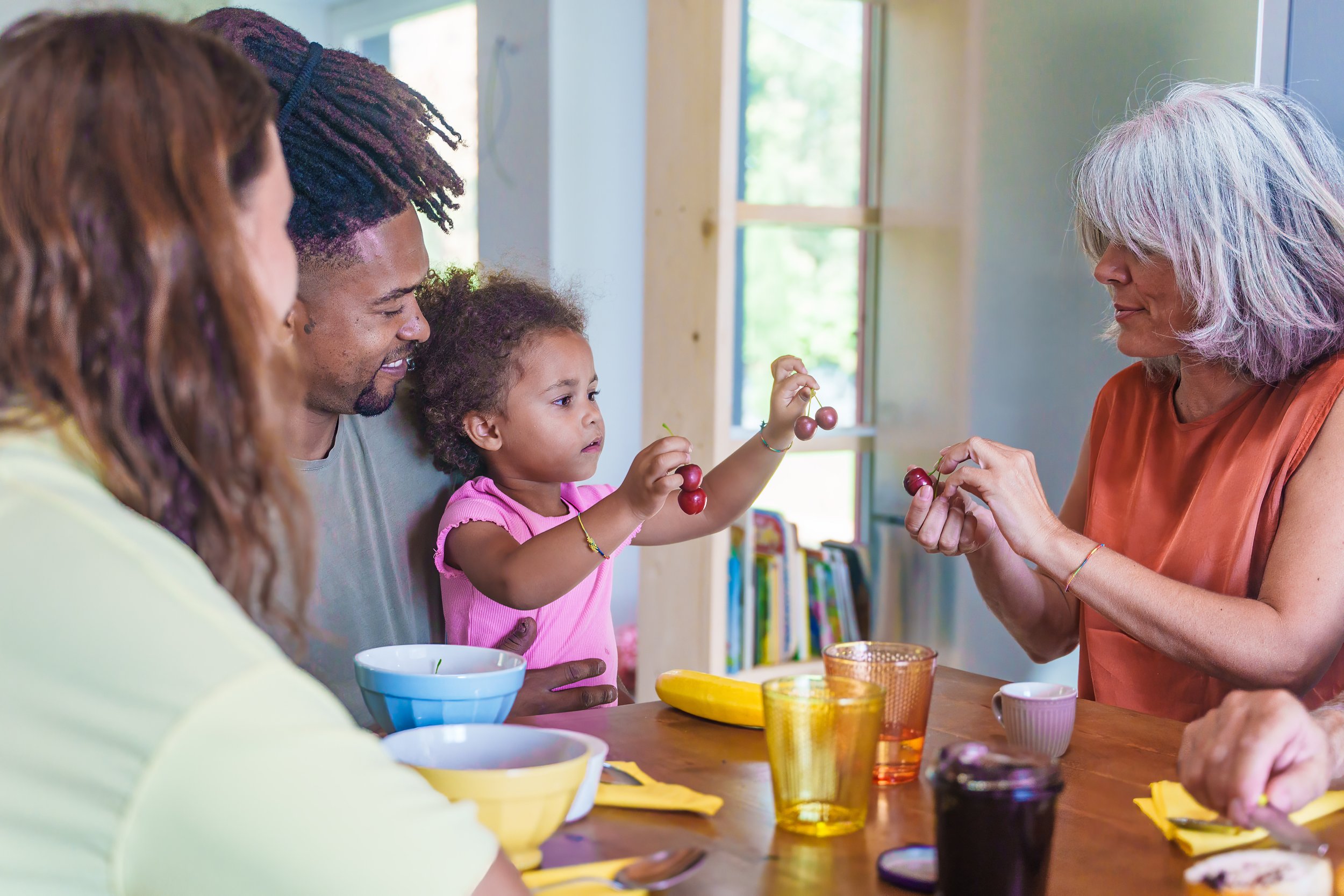 A multigenerational family at a dining table with colorful bowls and glasses, sharing grapes, in a bright room with natural light.