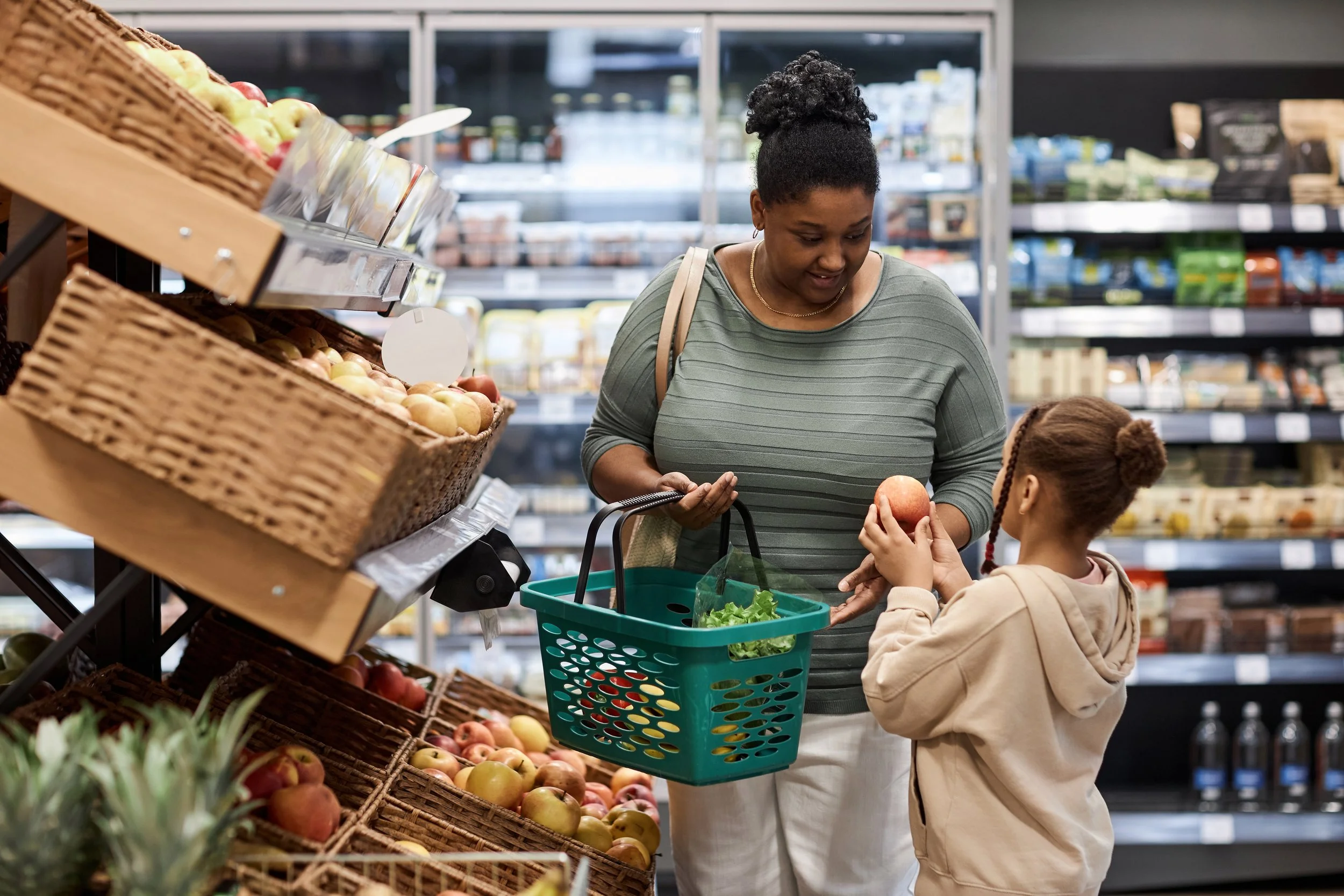 A woman and a young girl shopping for apples in a grocery store produce section. The woman is holding a shopping basket and looking at the young girl, who is handing her an apple.