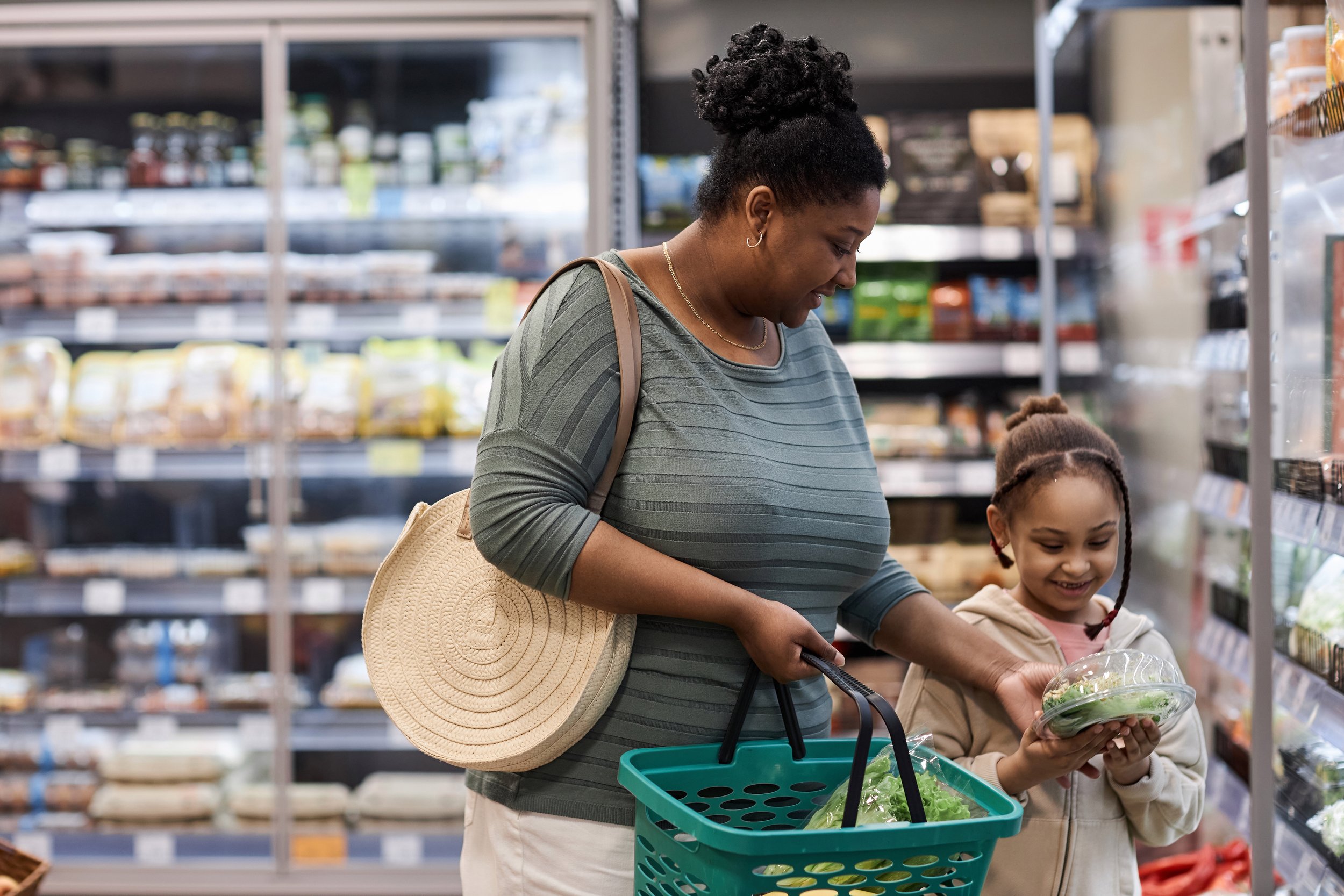 A woman and young girl shopping for produce in a grocery store, holding plastic containers with greens. The woman has a circular woven bag over her shoulder and is holding a teal shopping basket. The girl is smiling and looking at the greens.