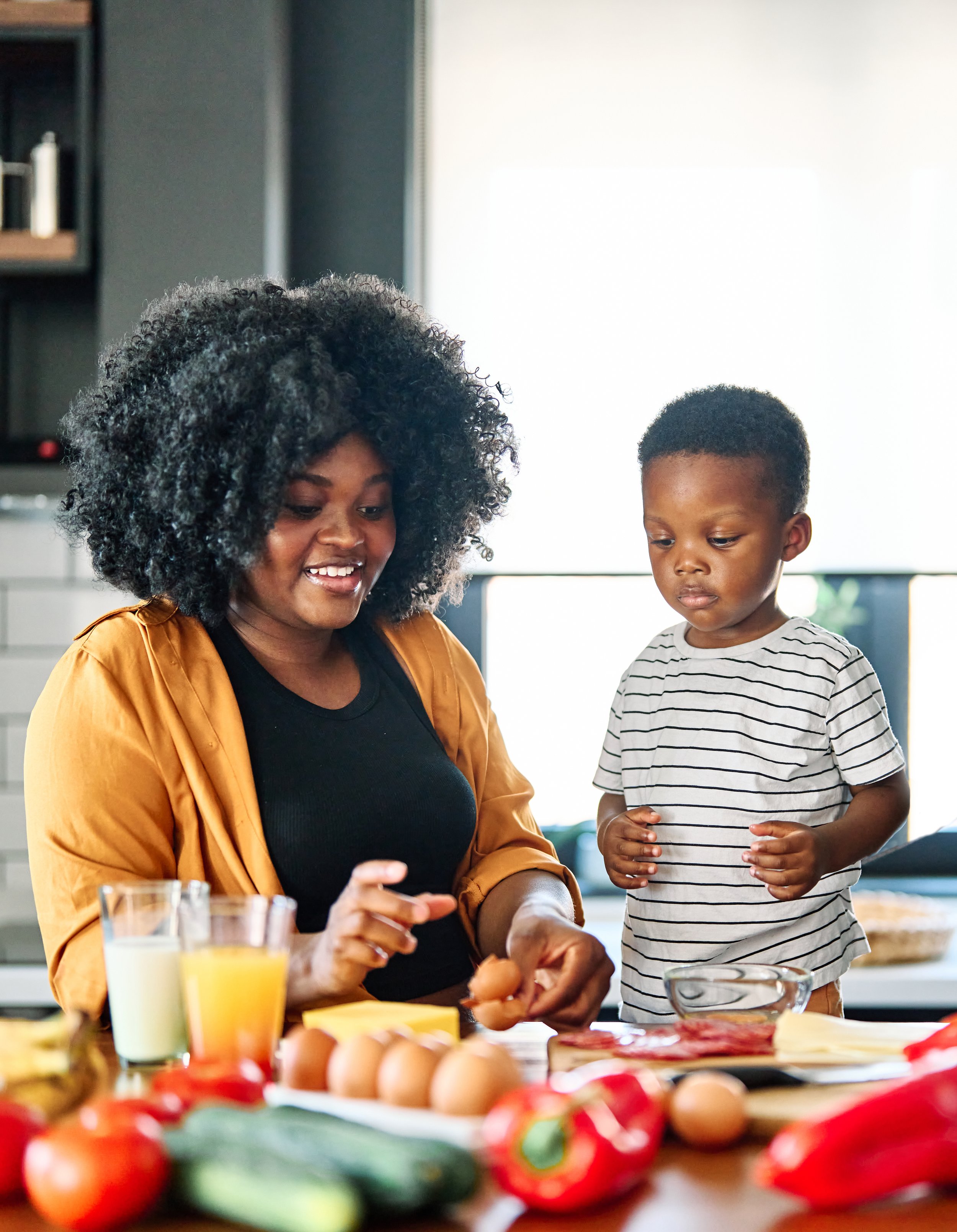 A woman and a young boy at a kitchen counter preparing food with eggs, vegetables, and drinks.
