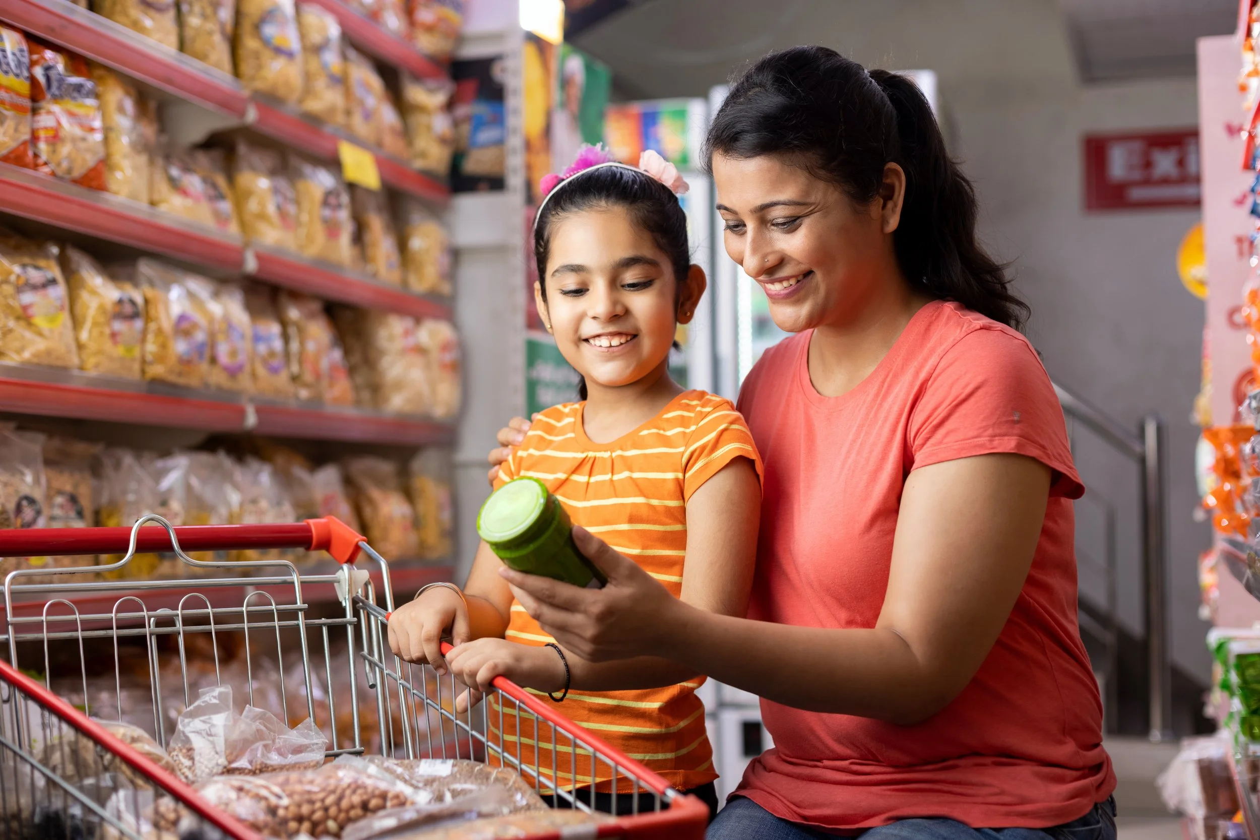 A woman and a young girl shopping for snacks at a grocery store, looking at a jar the girl is holding.