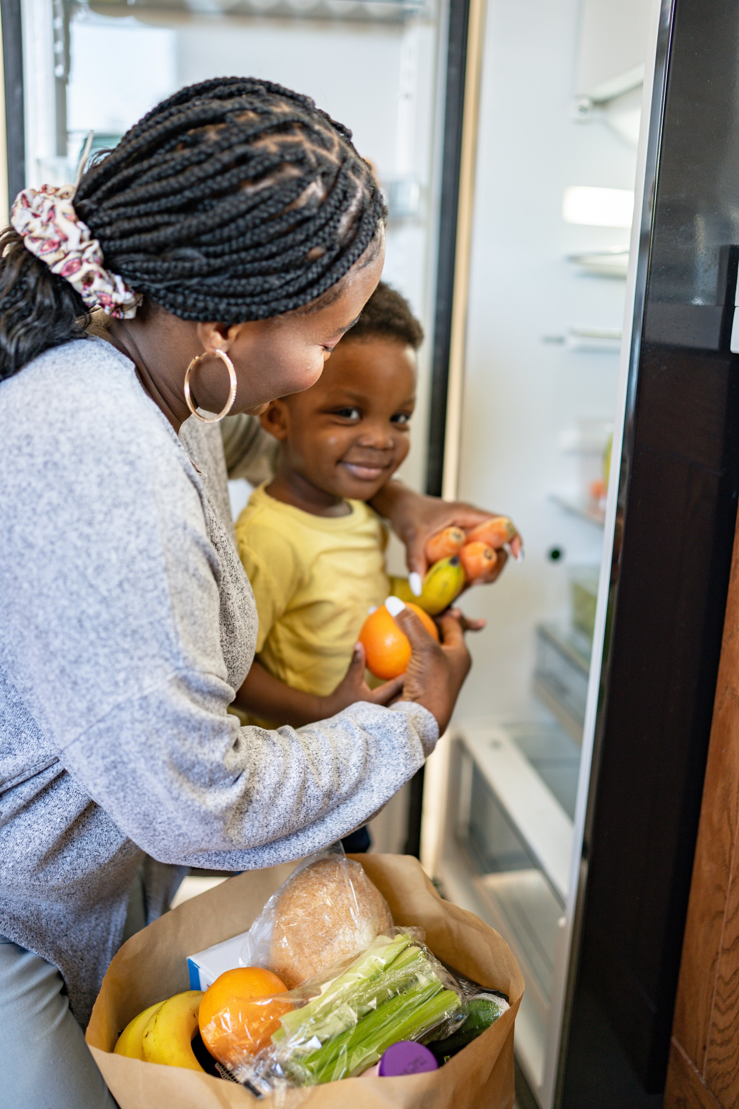 A woman and a young boy looking into an open refrigerator, holding vegetables and fruits, with a grocery bag of produce in front of them.