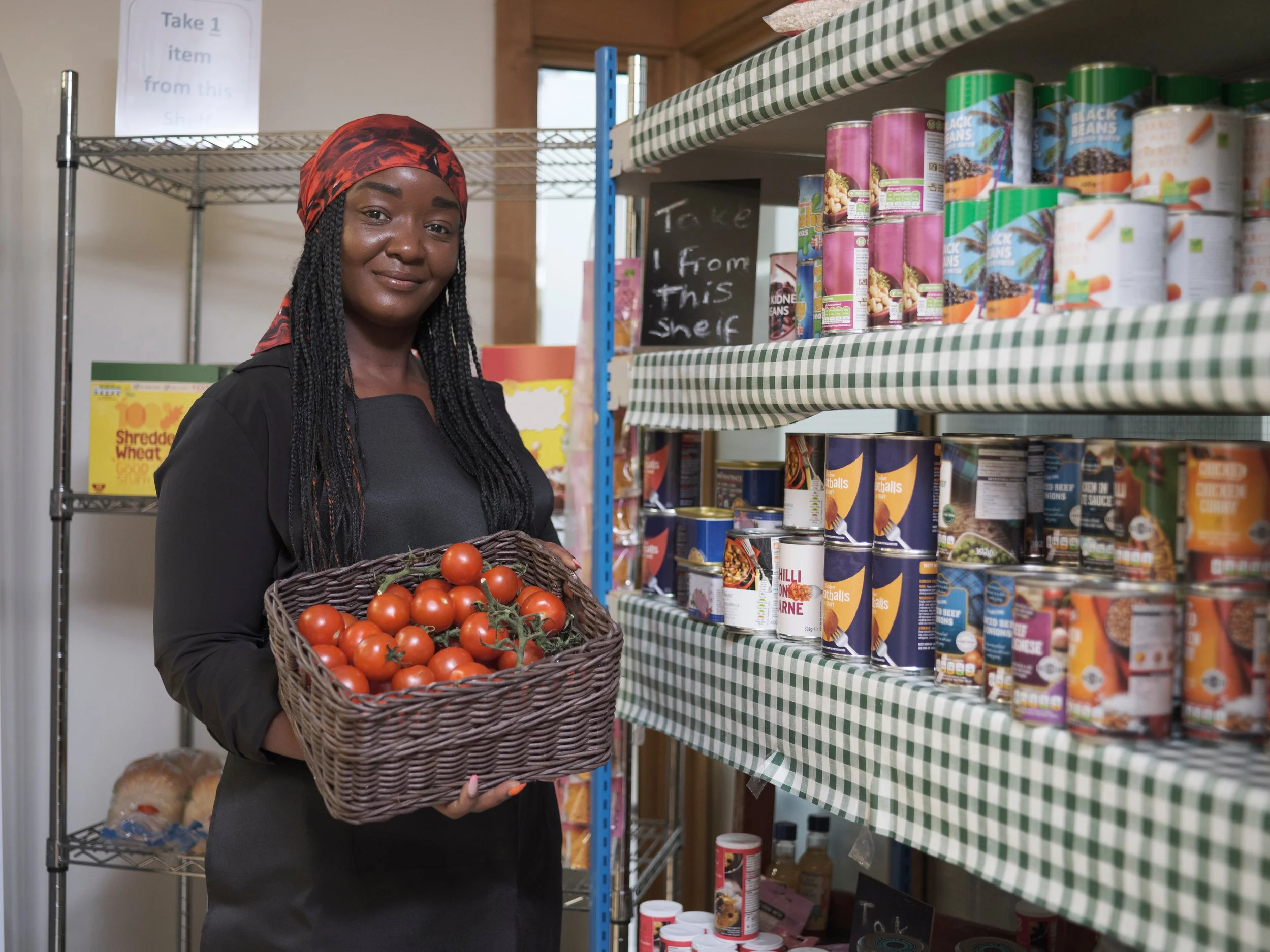 A woman wearing a black top and red headscarf holding a basket of fresh tomatoes in a grocery store aisle with shelves of canned goods and a sign that says 'Take 1 from this shelf'.