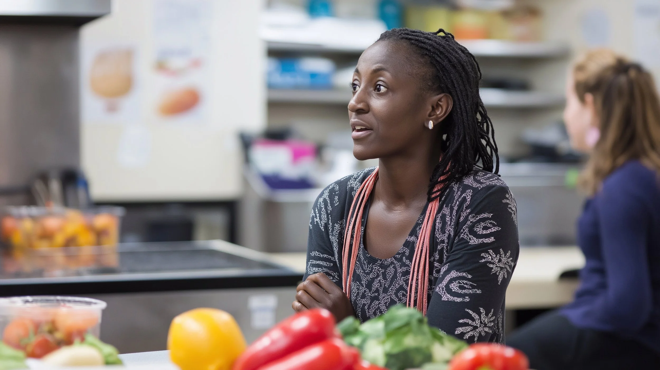 A woman with dark skin and dreadlocks sitting at a table in a kitchen or food preparation area, surrounded by various vegetables and fruits, engaging in conversation.