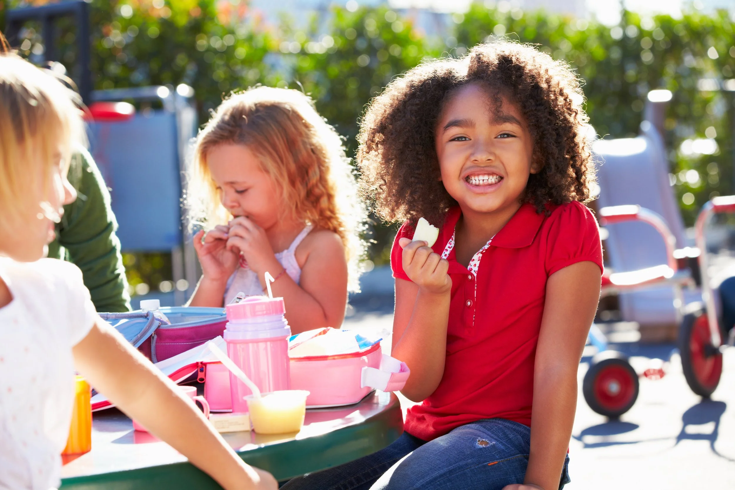 Young girl in a red shirt smiling and eating a snack while sitting at a picnic table outdoors with other children.