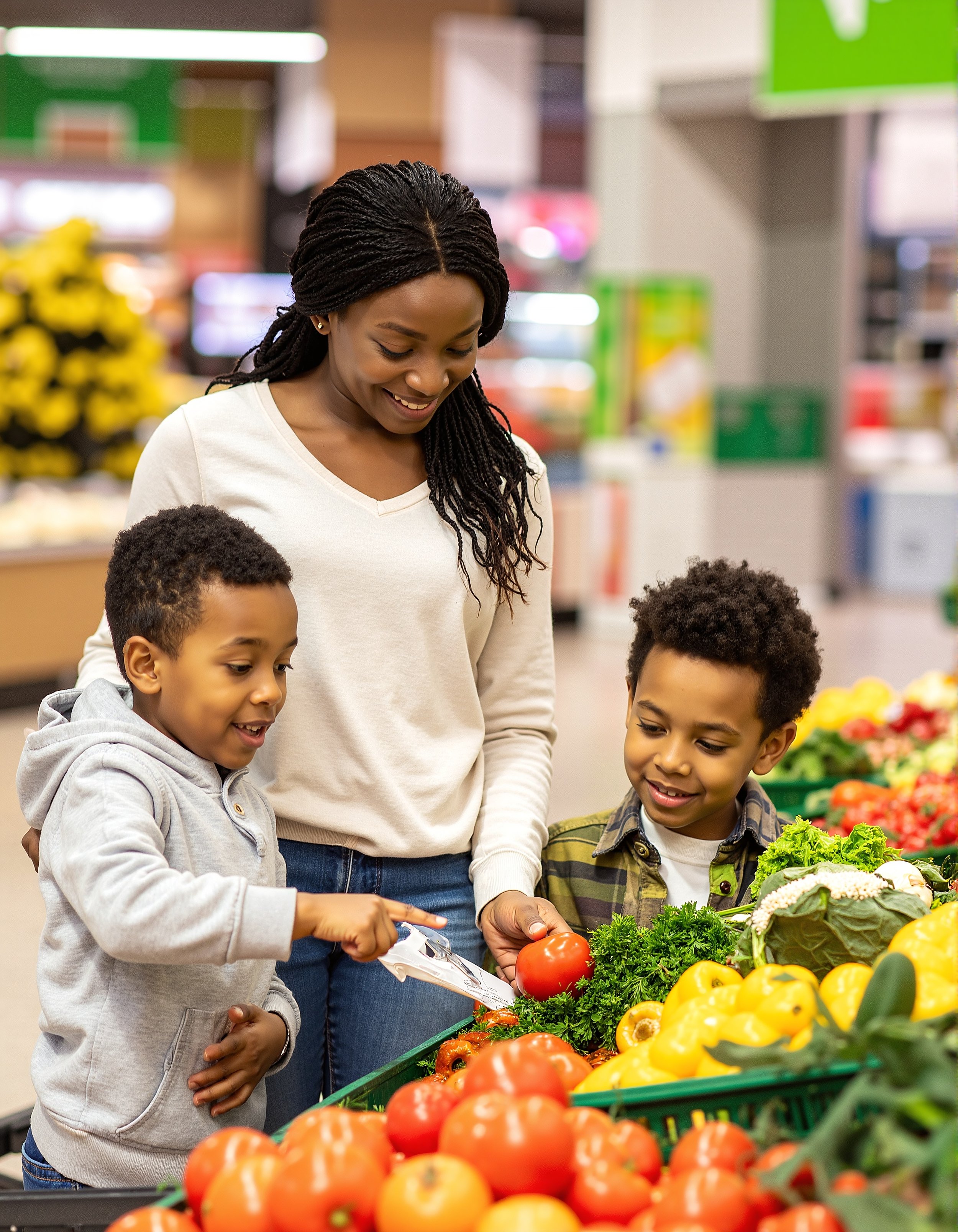 A woman with two young boys shopping for fresh vegetables in a grocery store, with tomatoes, yellow peppers, and lettuce visible.