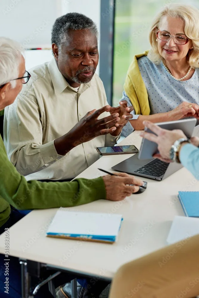 Group of diverse senior adults in a meeting, with laptops and notepads, engaging in discussion.
