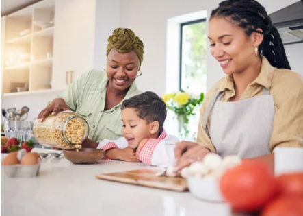 Three people in a kitchen, a woman pouring cereal for a smiling young boy, with another woman nearby.