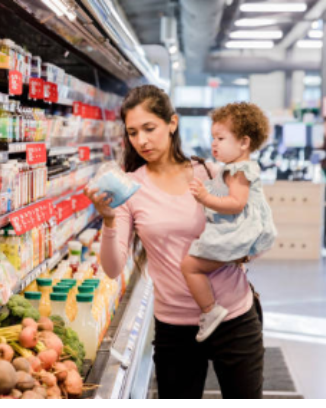 A woman shopping in a grocery store holding a young child on her hip. She is looking at a product in her hand while standing in front of refrigerated items and bottles of juice. The store has bright lighting and a modern layout.