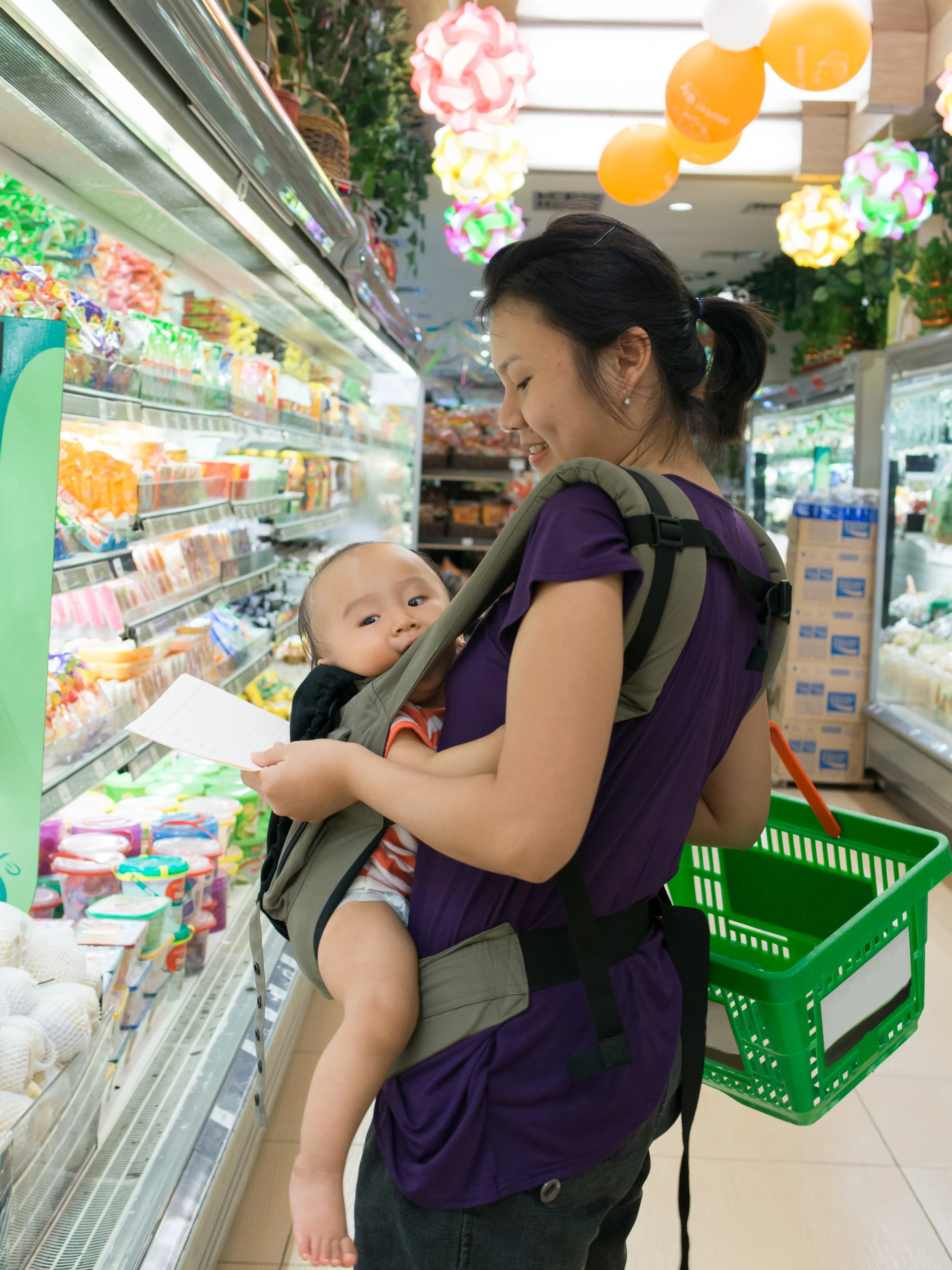 A woman carrying a baby in a front-facing baby carrier shopping in a grocery store, looking at products on the shelves, with colorful decorations hanging from the ceiling.