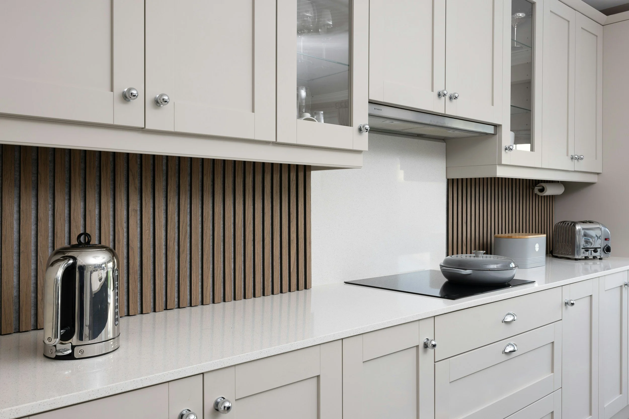 Kitchen counter with a stainless steel electric kettle, a rice cooker, a toaster, and a frying pan on an electric stovetop. White cabinets above and brown wood paneling in the background.
