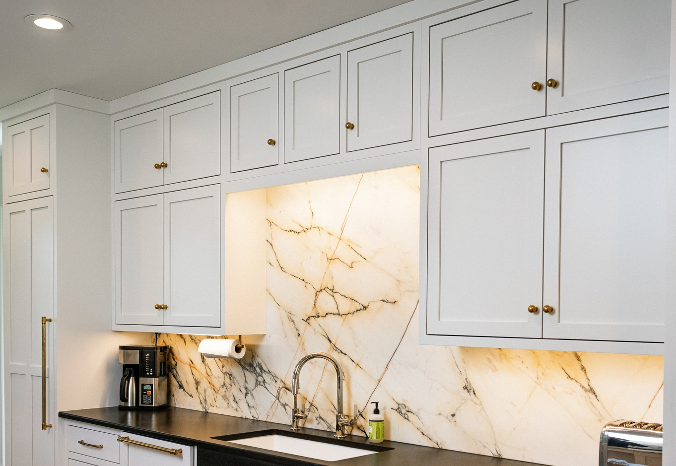Modern kitchen with white cabinets, marble backsplash, black countertop, coffee maker, soap dispenser, and gold hardware
