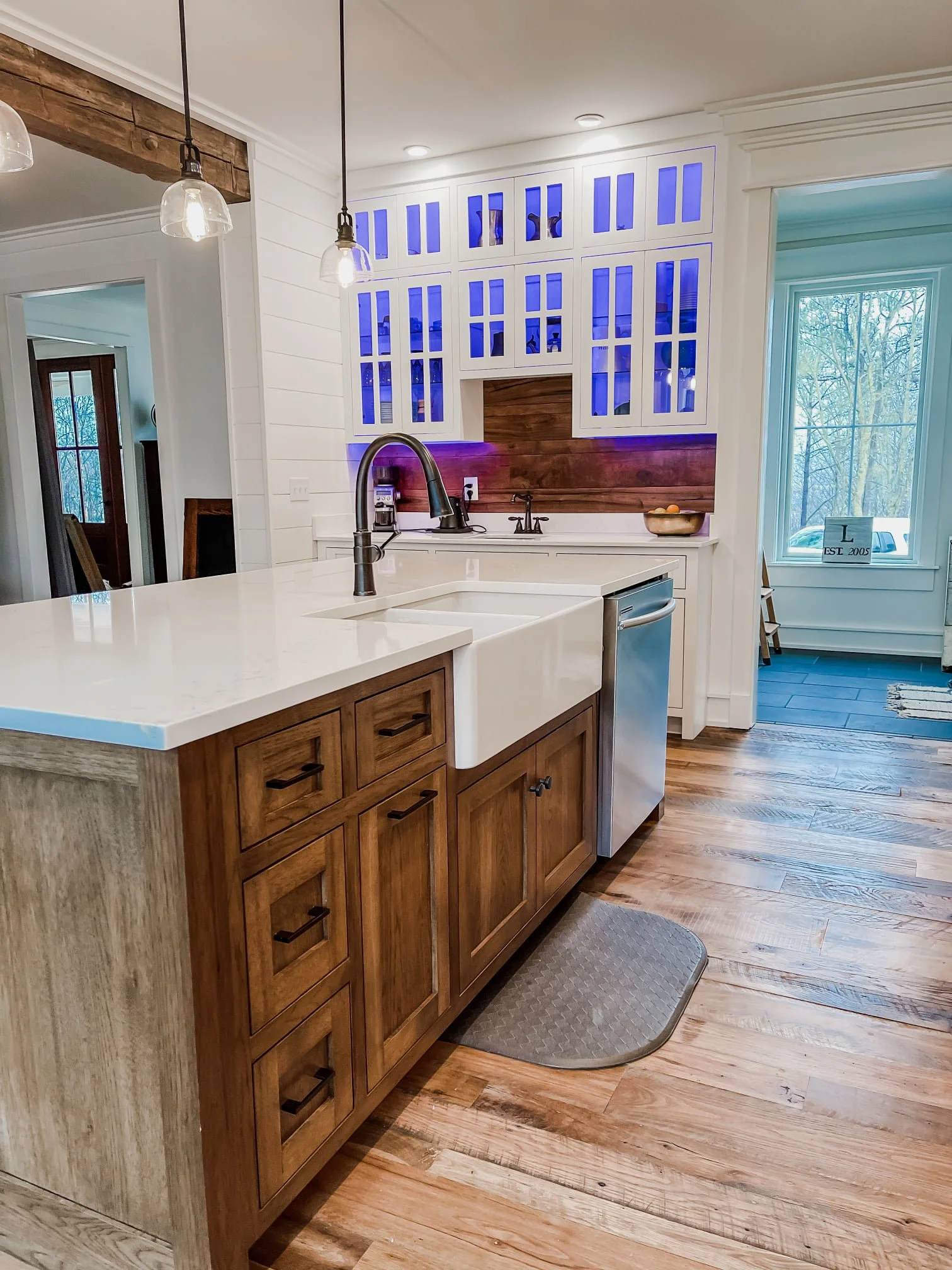 Modern kitchen with wooden cabinets, a white farmhouse sink, and pendant lights. A window with a view of trees outside and decorative wood paneling with purple lighting in the background.