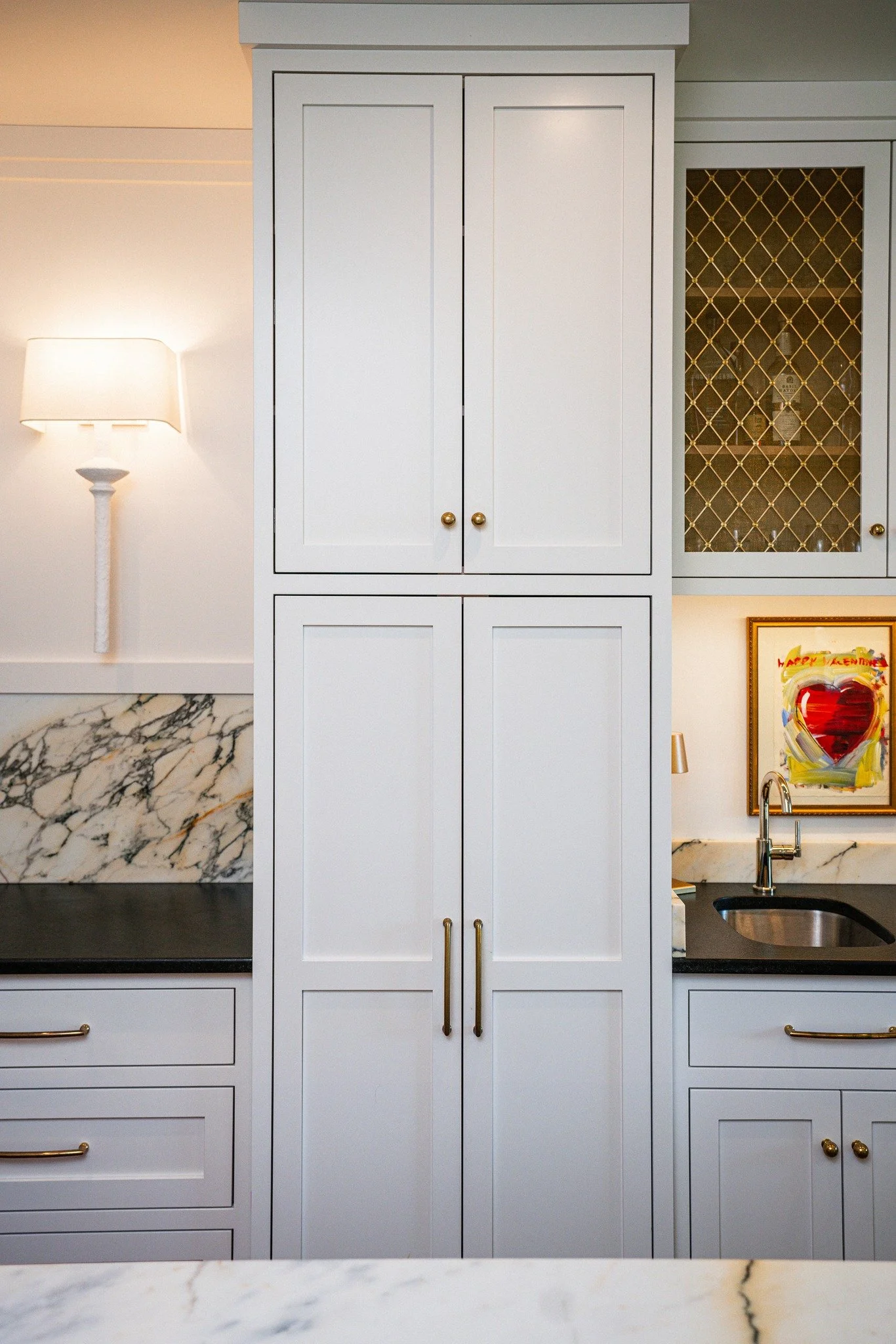 Kitchen with white cabinets, marble countertops and backsplash, black sink, brass handles, and a framed colorful art piece adorned with a heart.