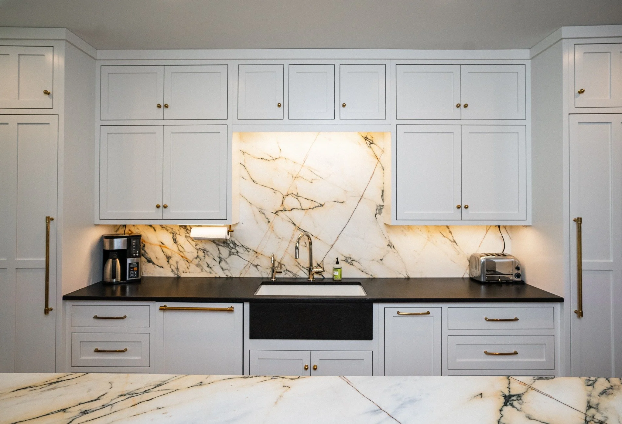 Kitchen with white cabinets, black countertop, marble backsplash, stainless steel appliances including a coffee maker and toaster, and a single sink with a gold faucet.