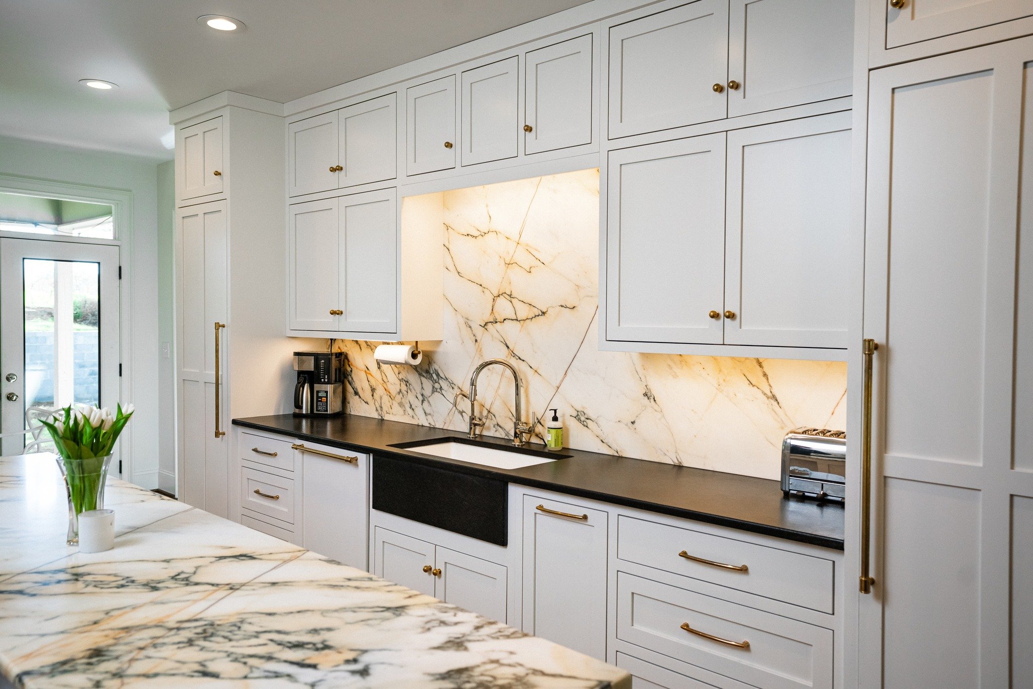 Modern kitchen with white cabinets, black countertop, marble backsplash, and gold hardware. A vase of tulips is on a marble island, with a coffee maker and toaster on the counter.
