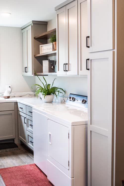 A laundry room with light gray cabinets, a white countertop, and a white washing machine. A potted plant is on the counter, and open shelves hold decor and a small speaker.