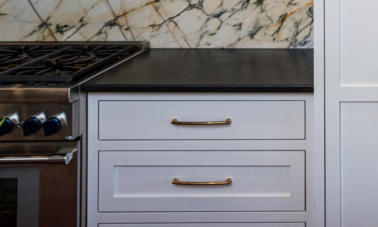 Close-up of a kitchen countertop, with a marble backsplash and white cabinets with gold handles, partially showing a stainless steel oven on the left.