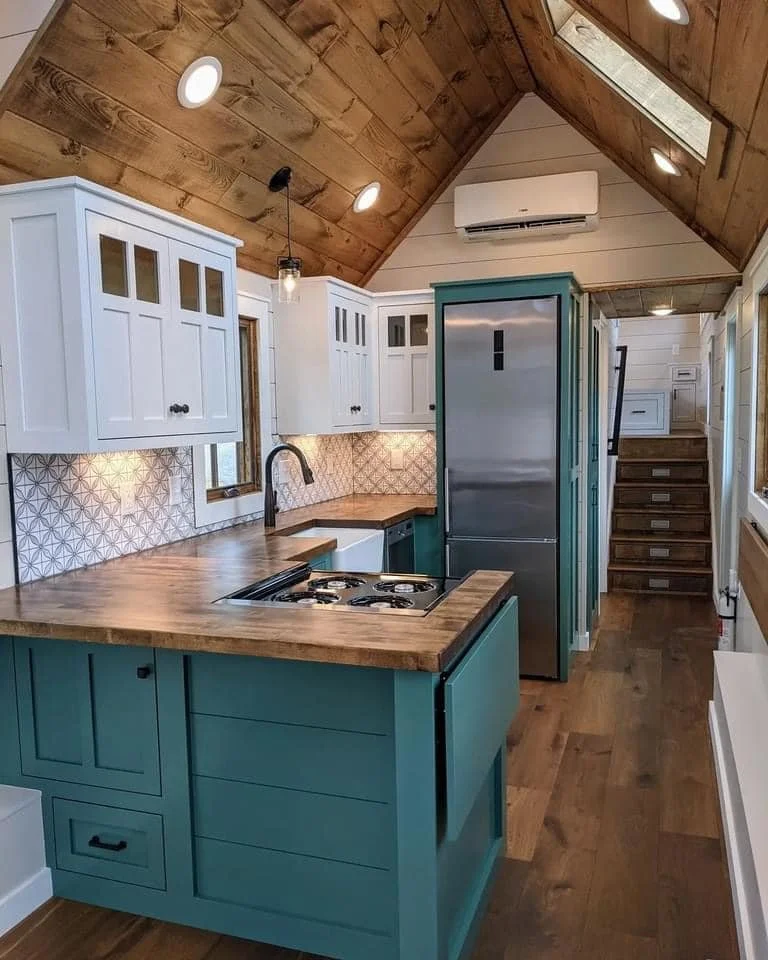 Kitchen with wooden ceiling, white upper cabinets, teal lower cabinets, patterned backsplash, small window, stainless steel refrigerator, staircase leading up, air conditioner mounted on wall, hanging pendant light.