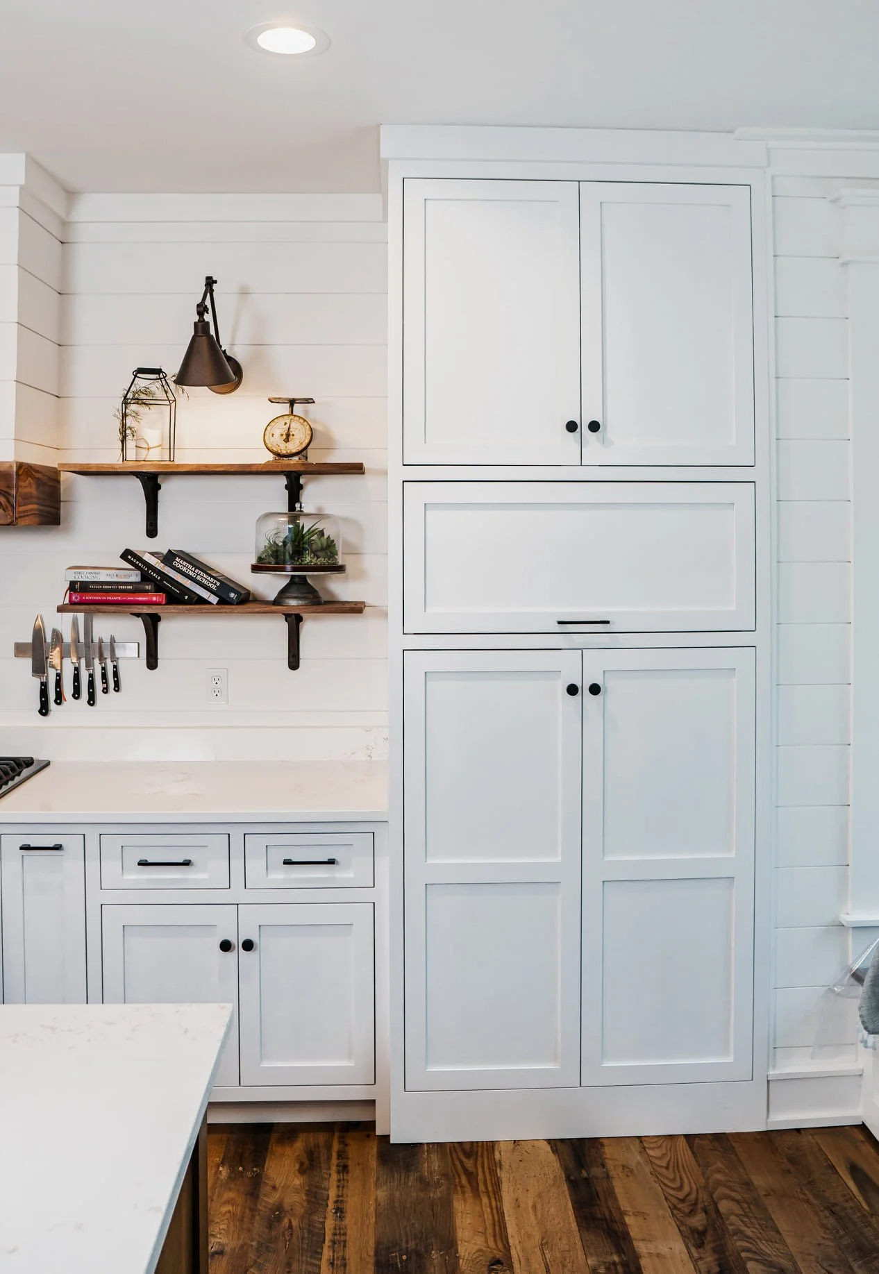 White kitchen cabinets and shelves with black handles, a wooden countertop, and a wooden floor. Shelves hold decorative items, books, a small scale, and utensils.