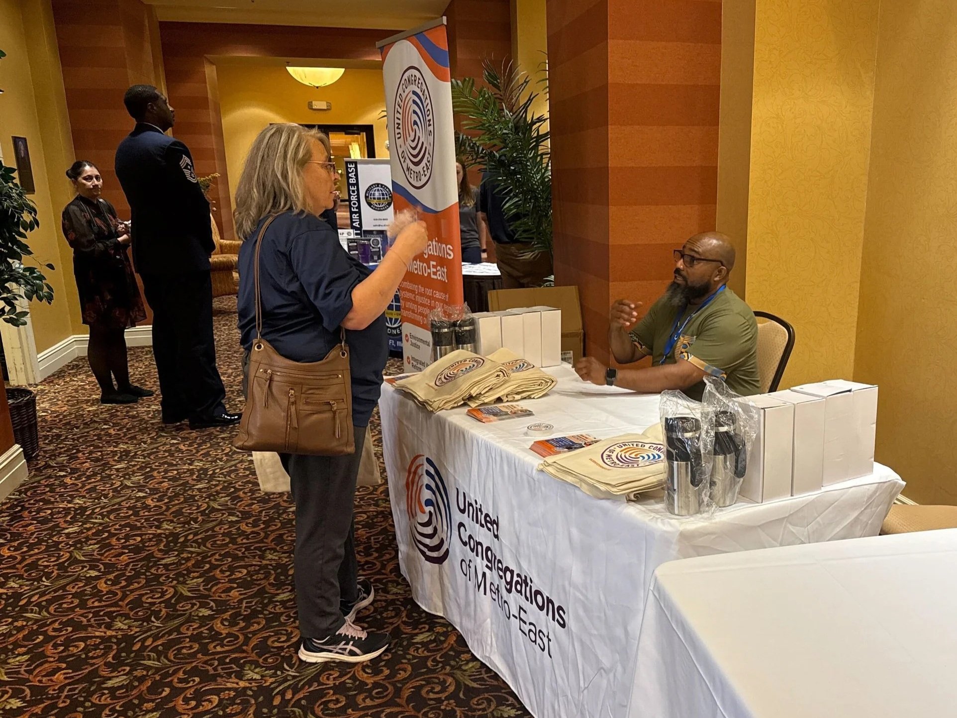 One woman standing talking with a man who is sitting. There is a table between them with a United Congregations of Metro-East table cloth and a banner behind them.