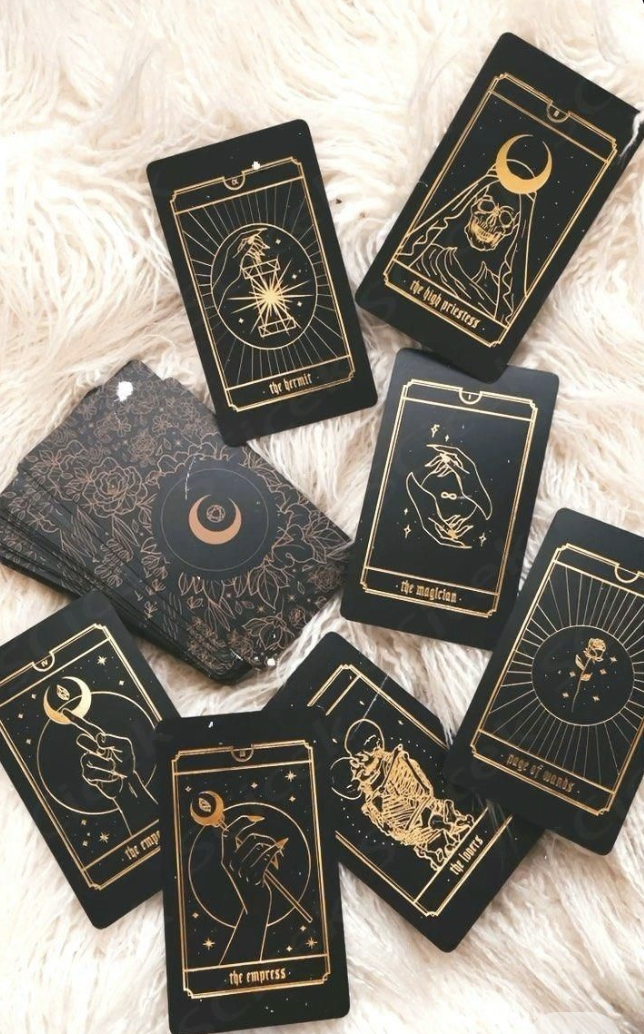 A spread of tarot cards with black backgrounds and gold illustrations, featuring various mystical symbols and titles like "the high priestess," "the empress," and "the magician," laid on a soft, cream-colored, fluffy surface.