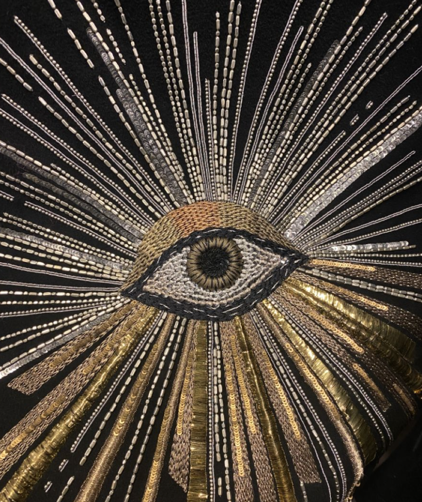 An embroidered eye design with radiating lines of beads and sequins in gold, silver, and cream colors, set against a black background.