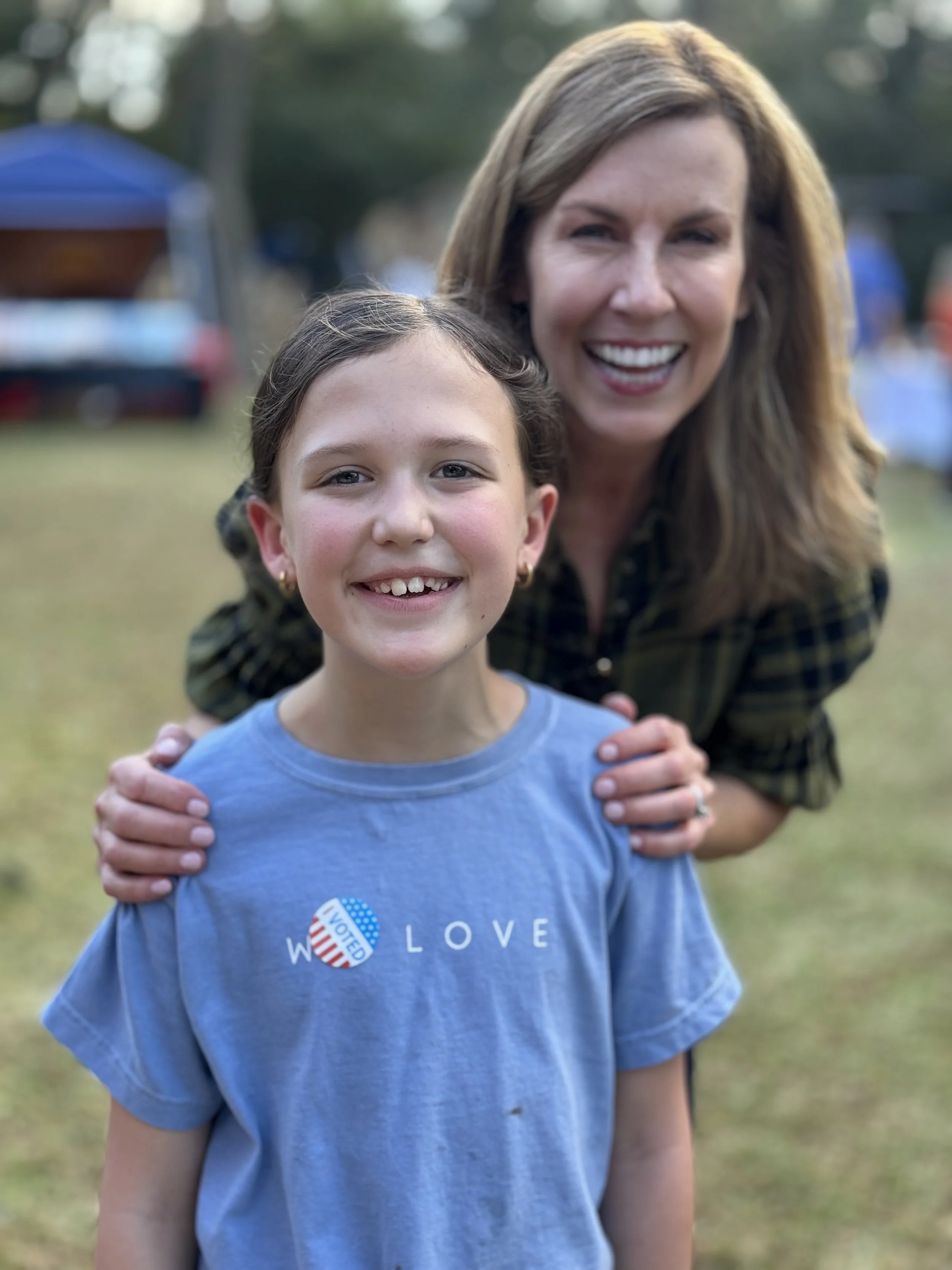 A young girl and a woman smiling outdoors, with the girl wearing a blue shirt that says 'LOVE' and a sticker that reads 'VOTE,' and the woman standing behind her with hands on her shoulders.